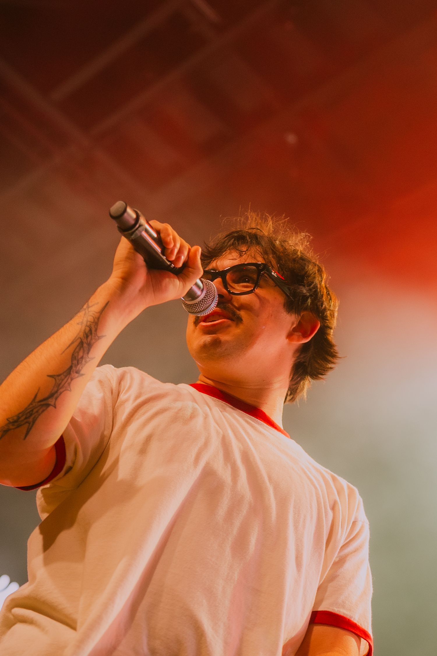 A performer in a white shirt sings passionately into a microphone against a red-tinted stage backdrop.