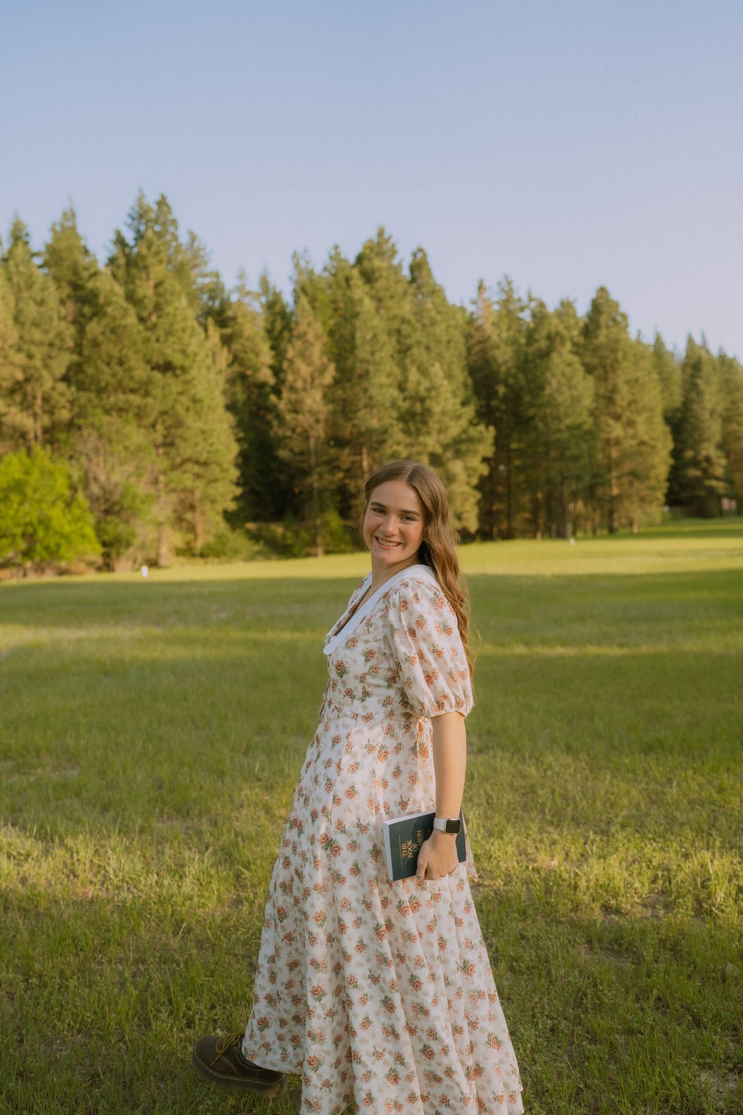 A floral maxi dress is worn in a sunny meadow with evergreen trees in the background.