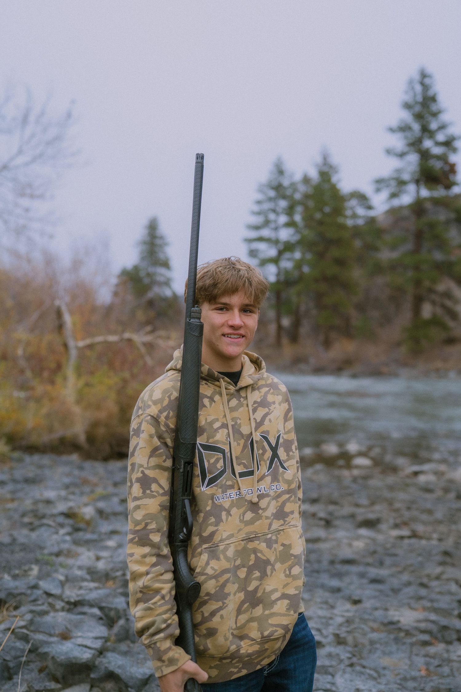 Person in camouflage hoodie holding hunting rifle stands by rocky riverbank with evergreen trees in background.