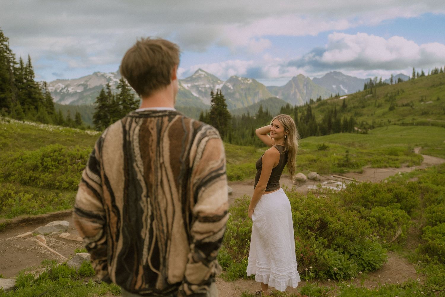 A couple walks hand in hand on a scenic mountain path with wildflowers and evergreens.