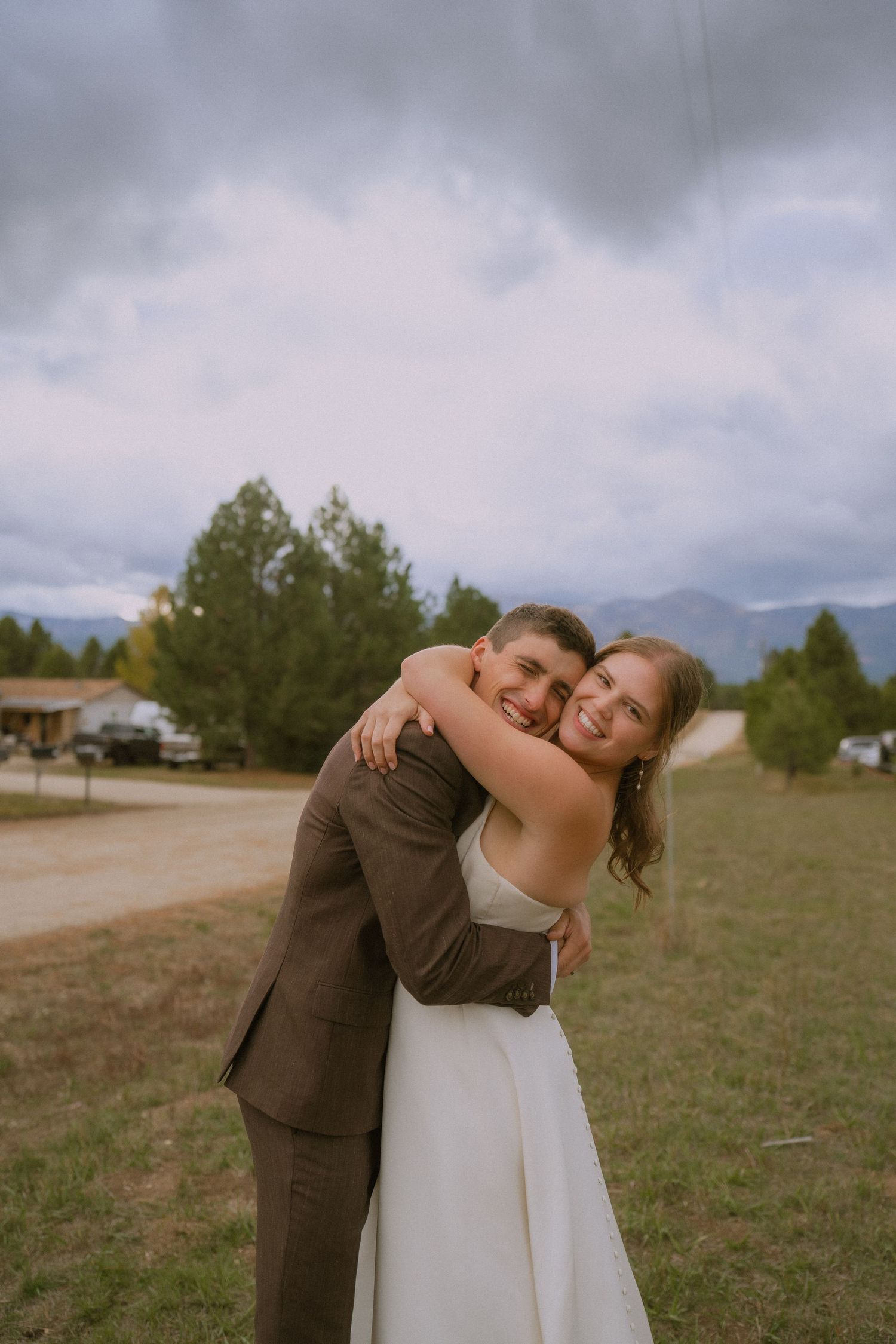 Two people embrace in a heartfelt hug during an outdoor wedding ceremony on a rural field with mountains in the background.
