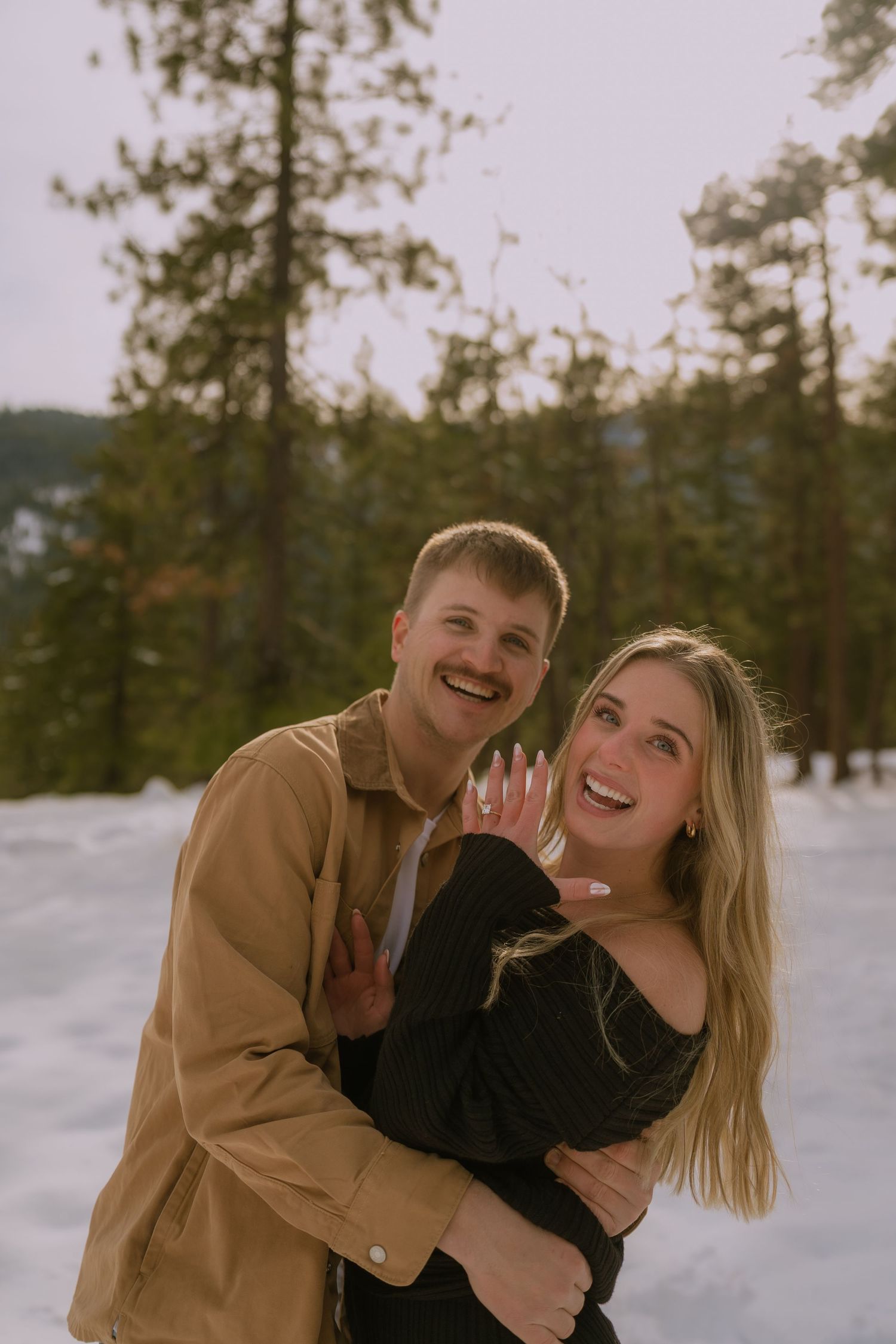 A couple laughs and embraces in a snowy winter landscape with evergreen trees in the background.
