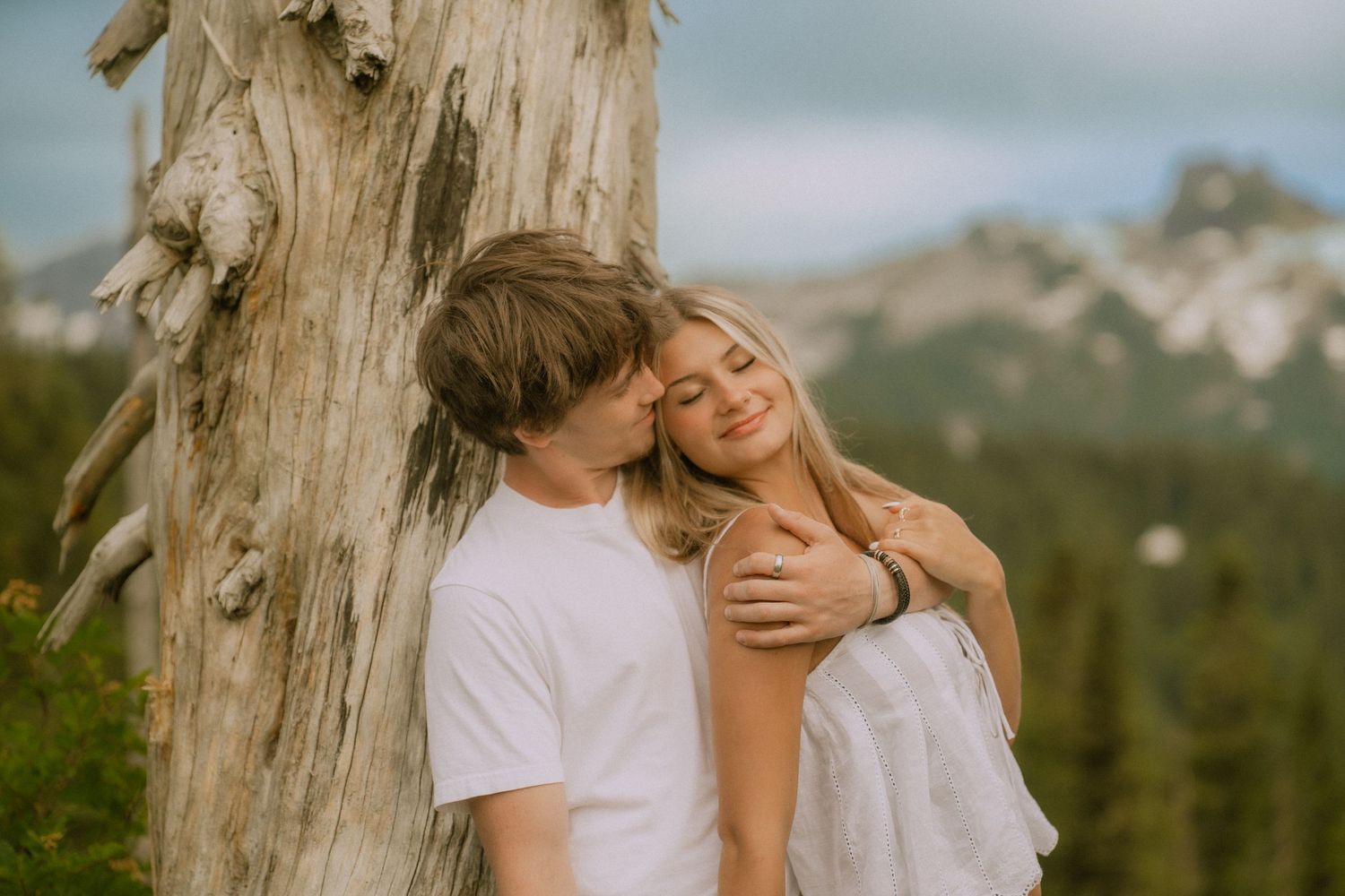 A romantic moment captured against a weathered tree trunk in a mountain setting.