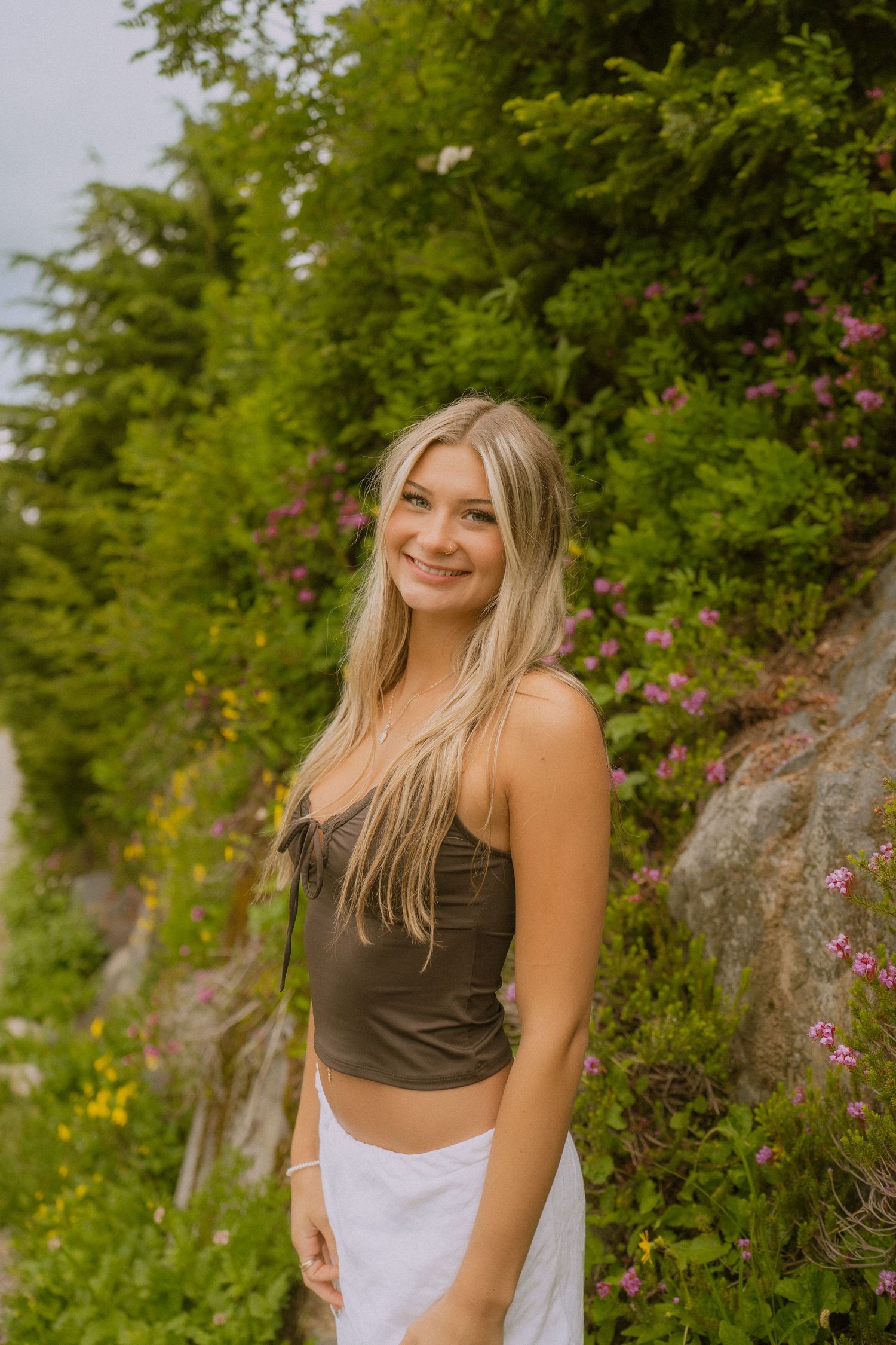 A person in casual summer attire poses outdoors against a backdrop of lush greenery and wildflowers.