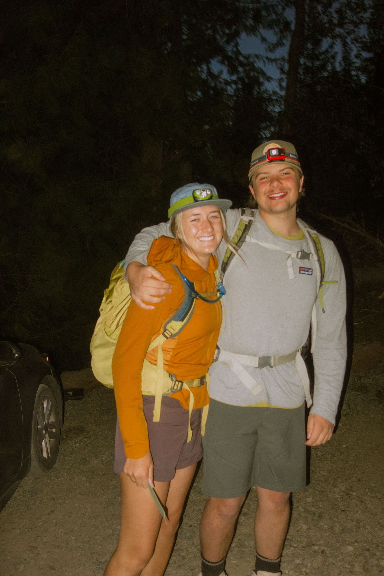 Two hikers wearing headlamps and backpacks stand together on a dark trail during a nighttime adventure.