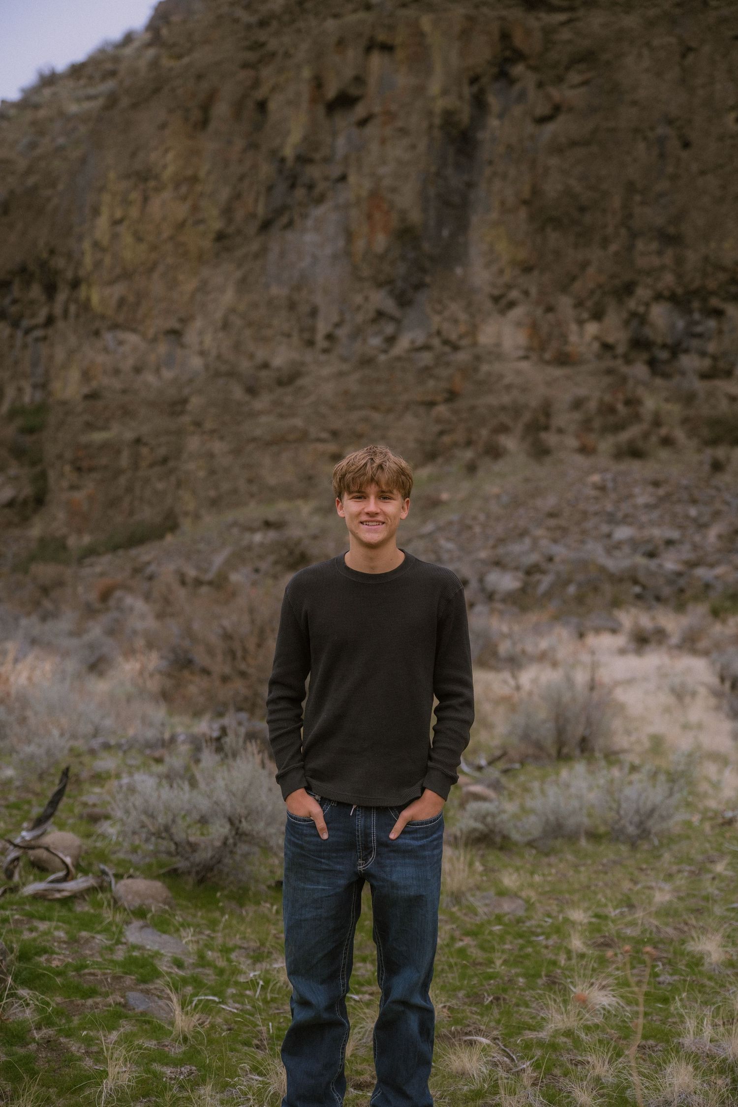 A person in a dark sweater and jeans stands outdoors against a rocky cliff backdrop in natural lighting.