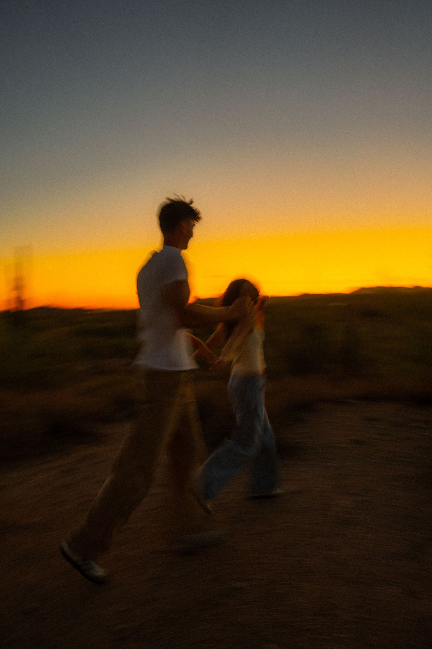 Two silhouetted figures walk across a desert landscape against a vibrant orange and yellow sunset sky.