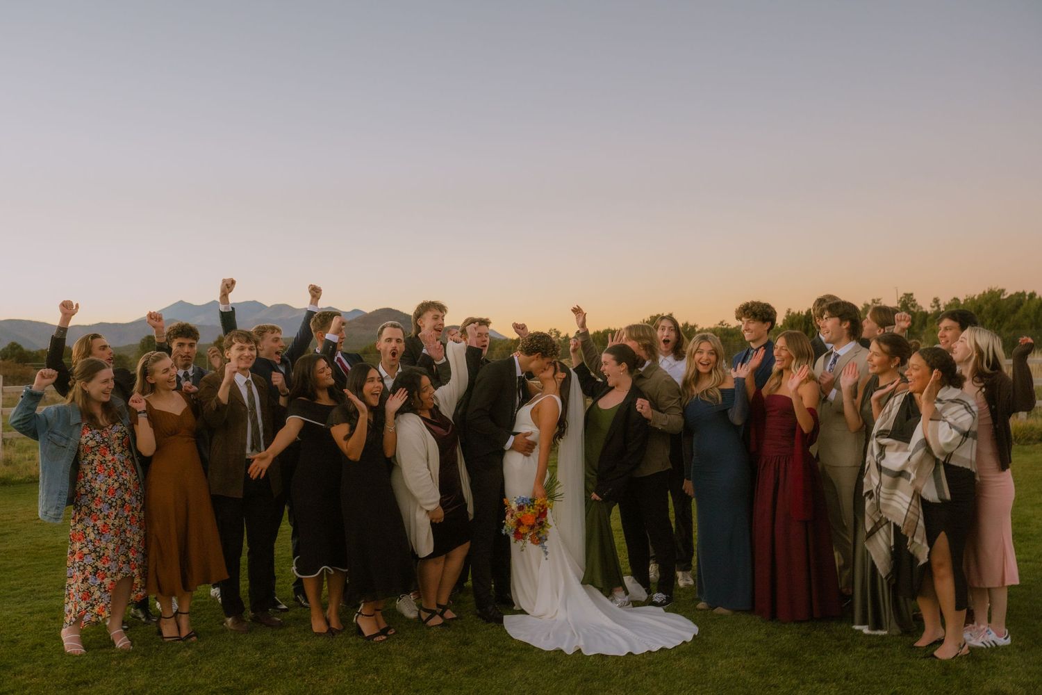 A large wedding party poses together for a panoramic group photo in a scenic outdoor setting.