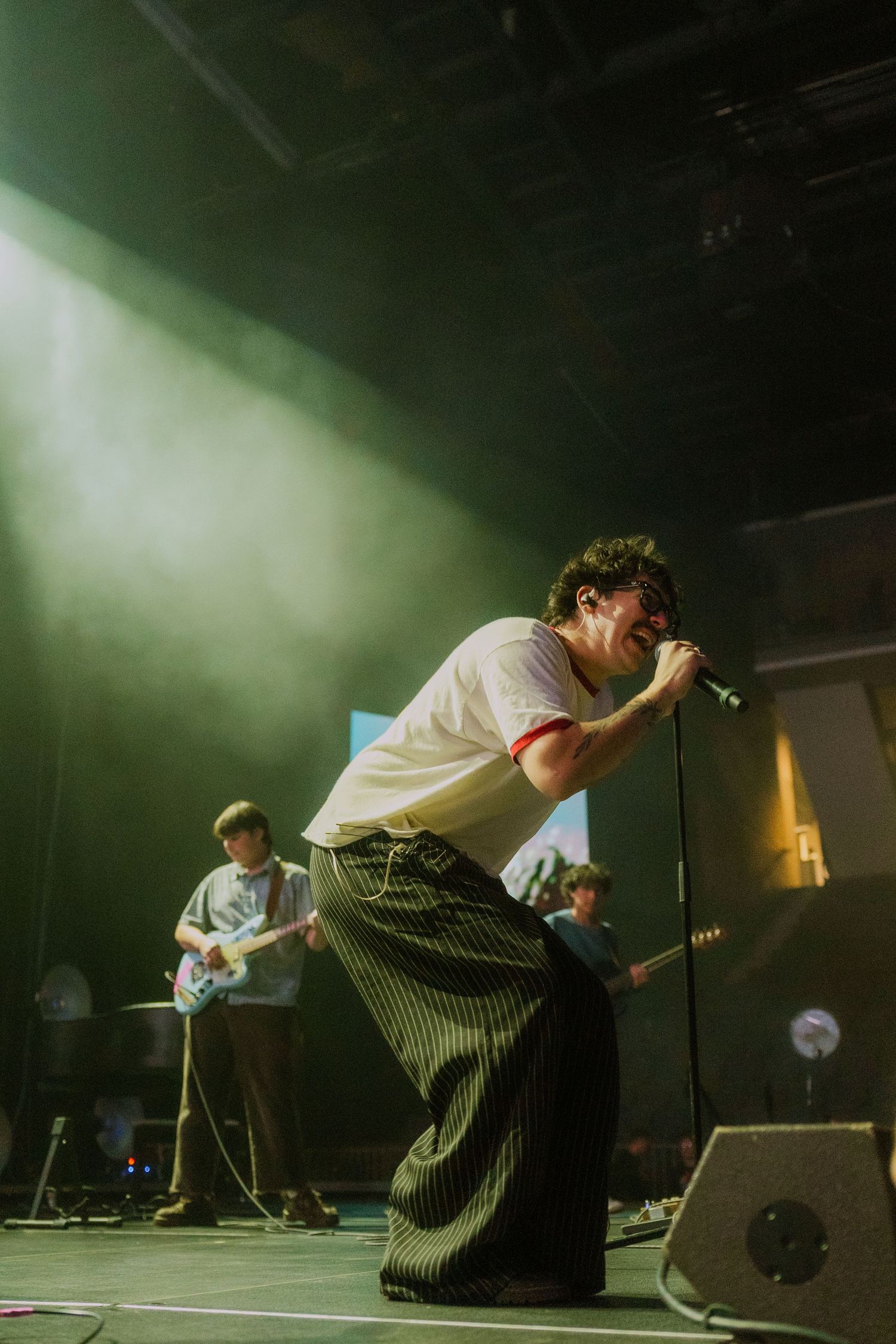 Energetic performer singing on stage in white shirt and striped pants with dramatic green lighting effects.