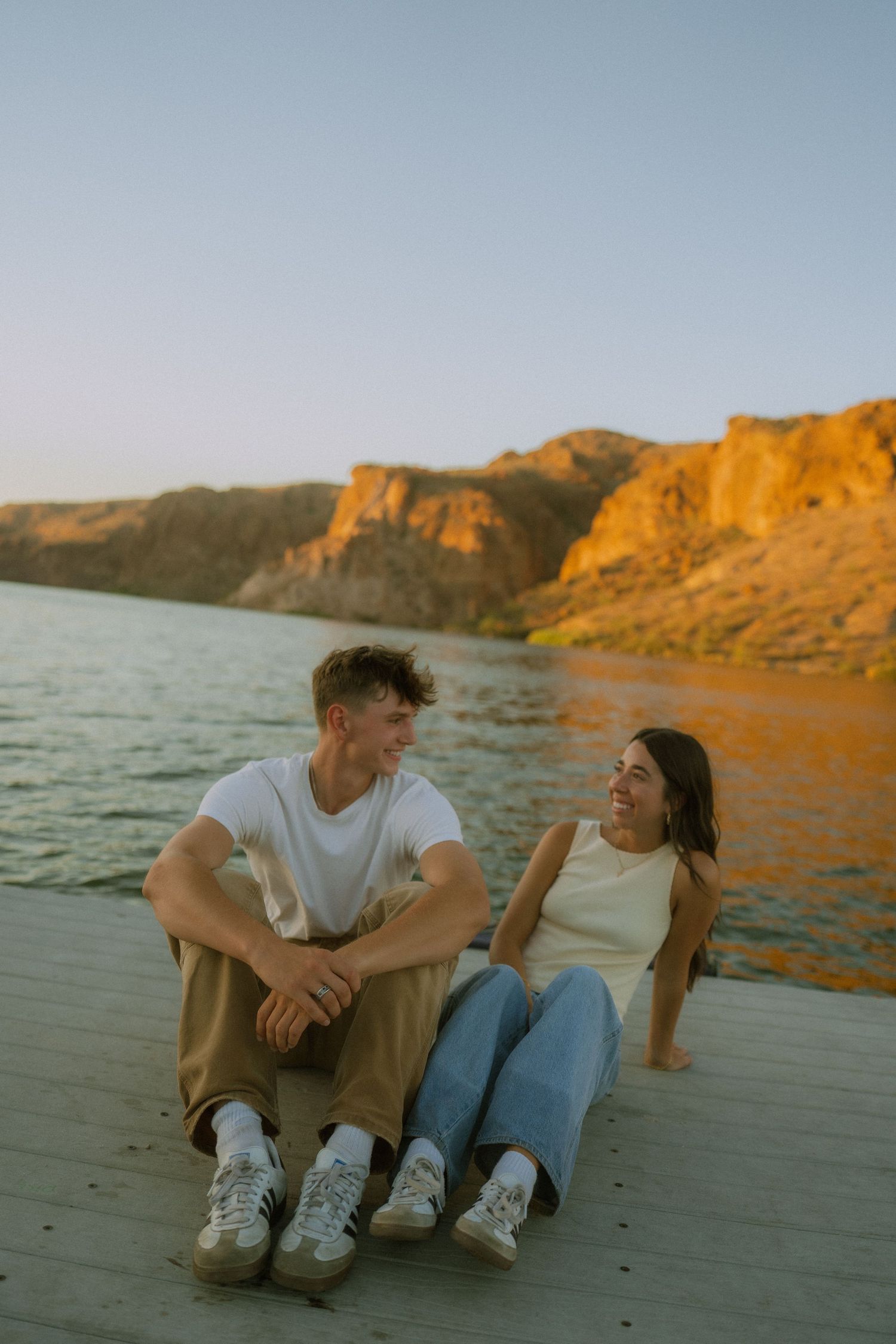 Two people sitting on a beach at sunset with golden cliffs and ocean in the background.