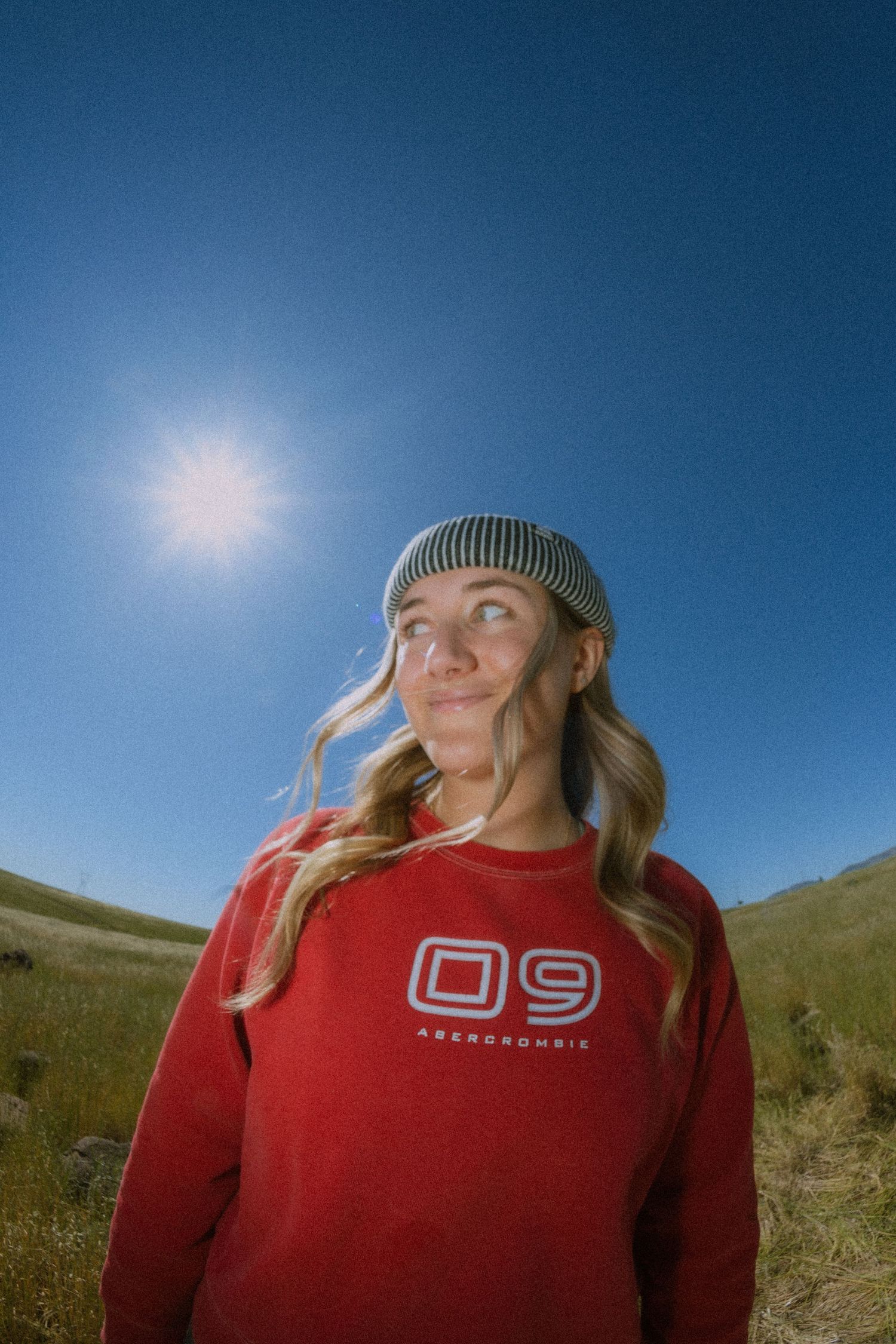A person in a red sweatshirt and knit beanie stands against a blue sky on a grassy hillside in a panoramic series.
