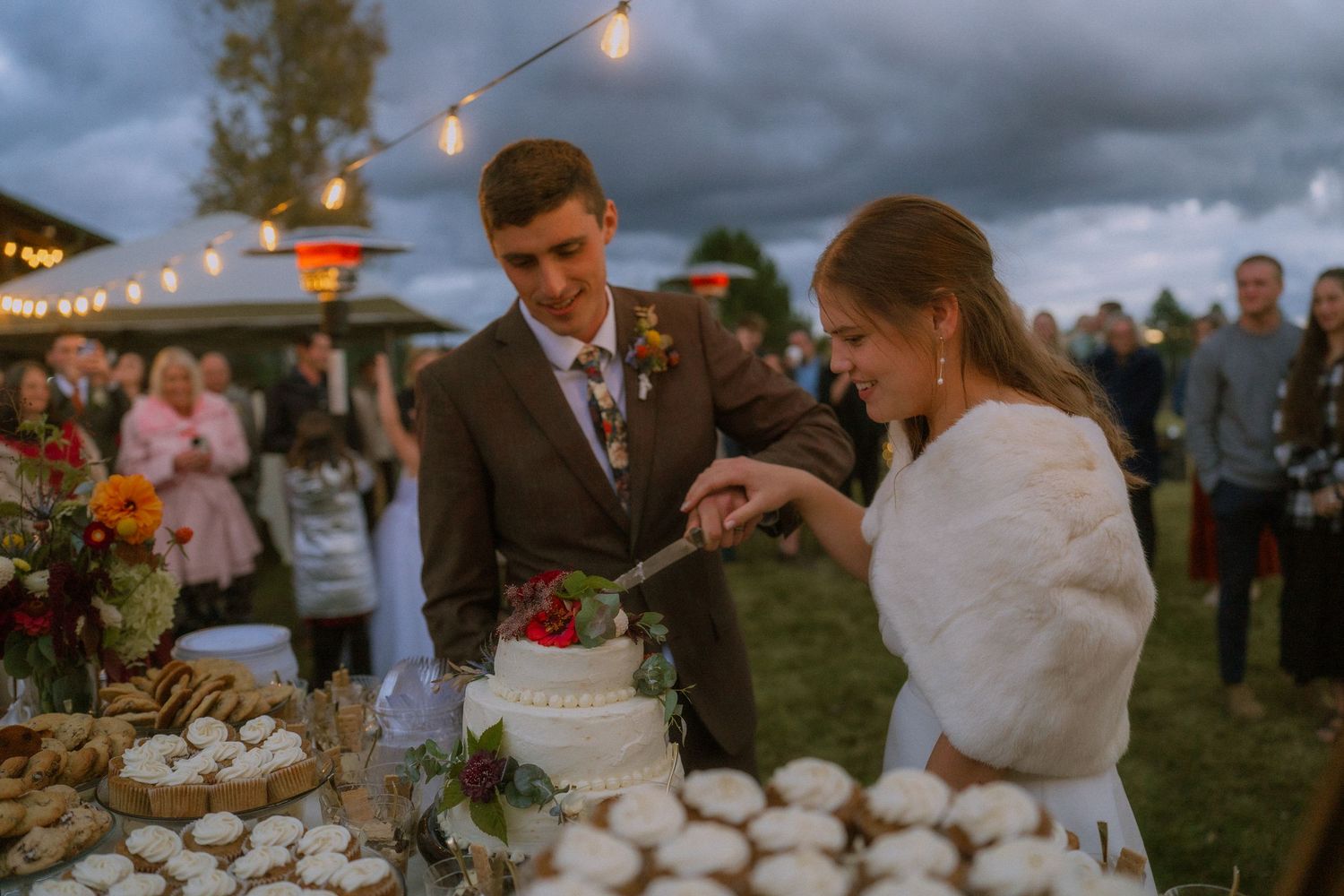 Wedding guests gather around a rustic outdoor table with string lights overhead during an evening celebration.
