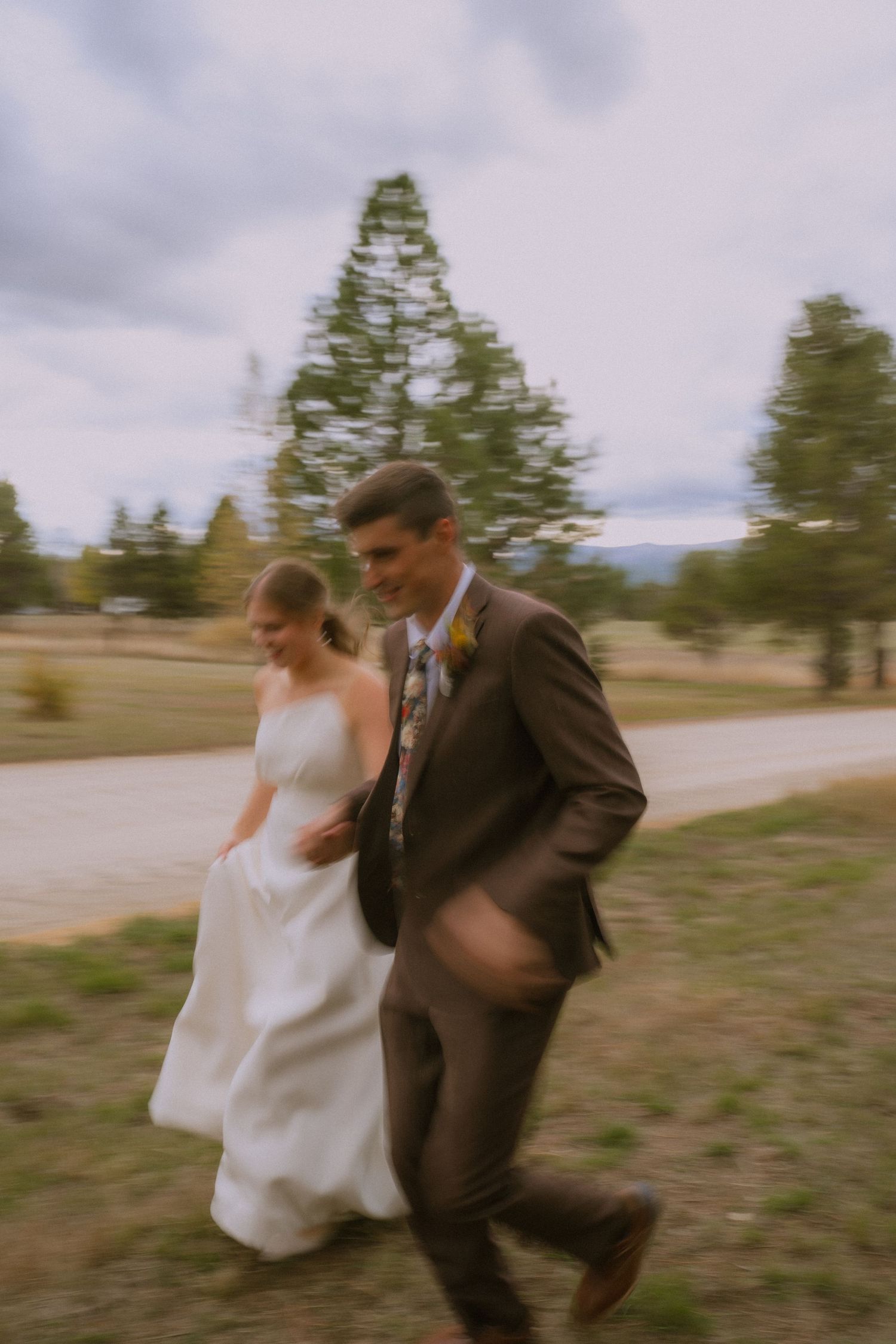 A couple in wedding attire running playfully across an outdoor venue with evergreen trees in the background.