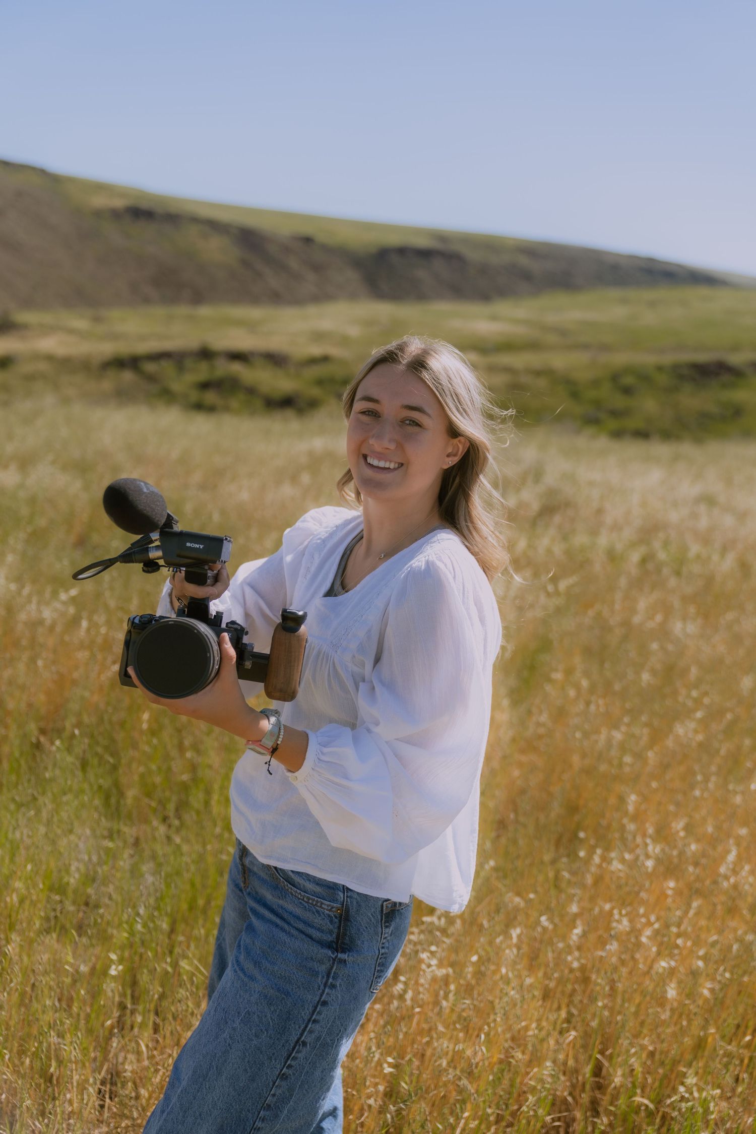 Videographer in white shirt and jeans films in golden grass field on sunny day with professional camera equipment.