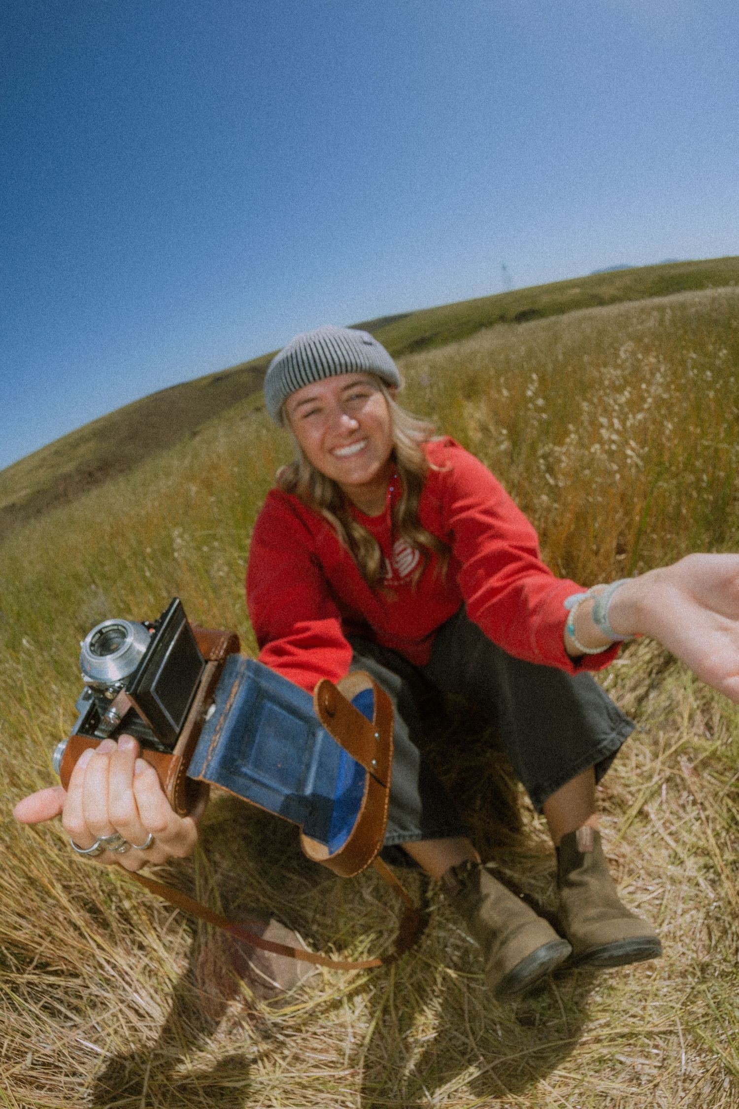 Person in red jacket lying in dry grass field taking photos with vintage camera on sunny day.