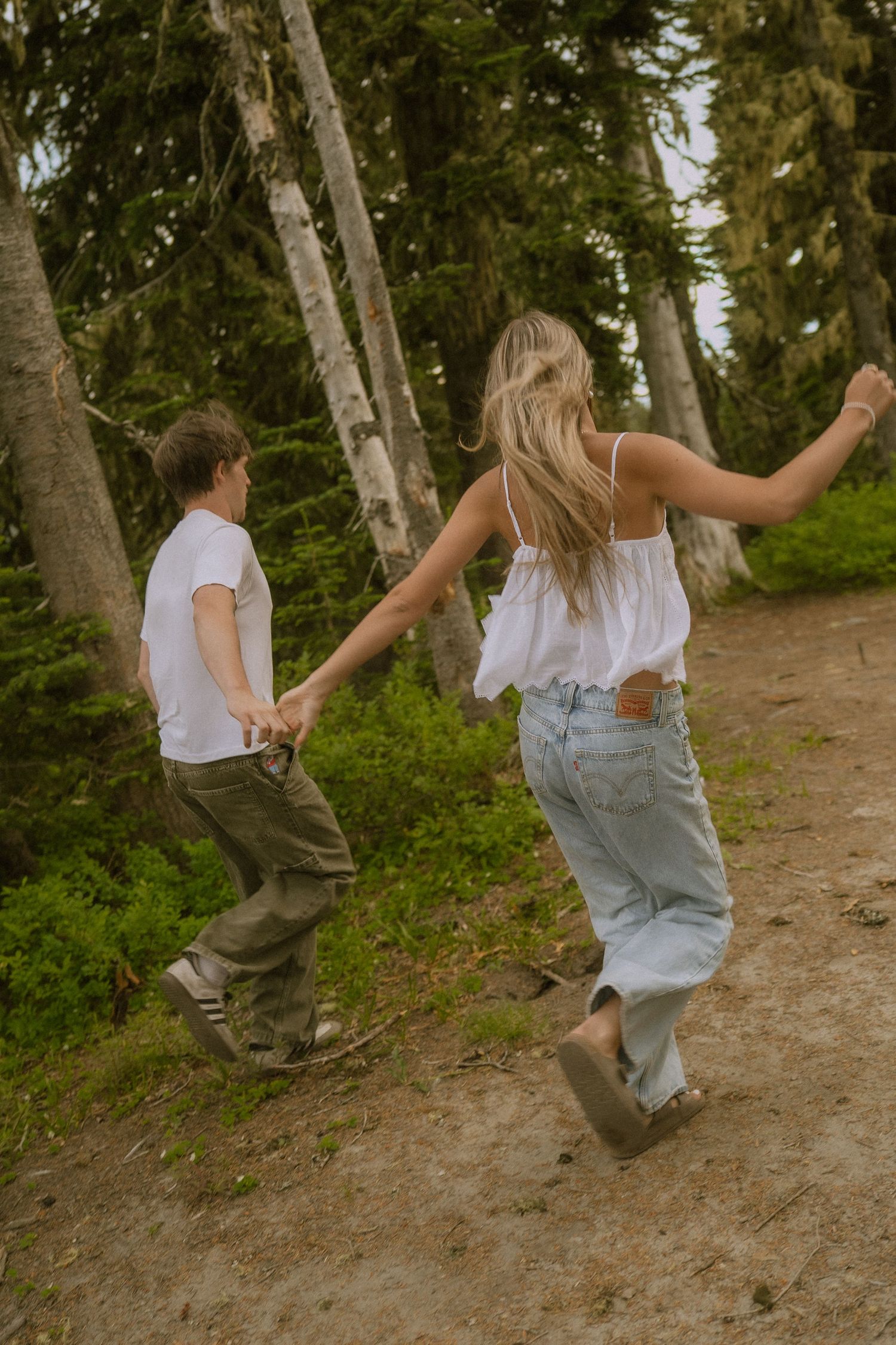 Two people dancing and spinning on a dirt trail through a forest of tall evergreen trees.
