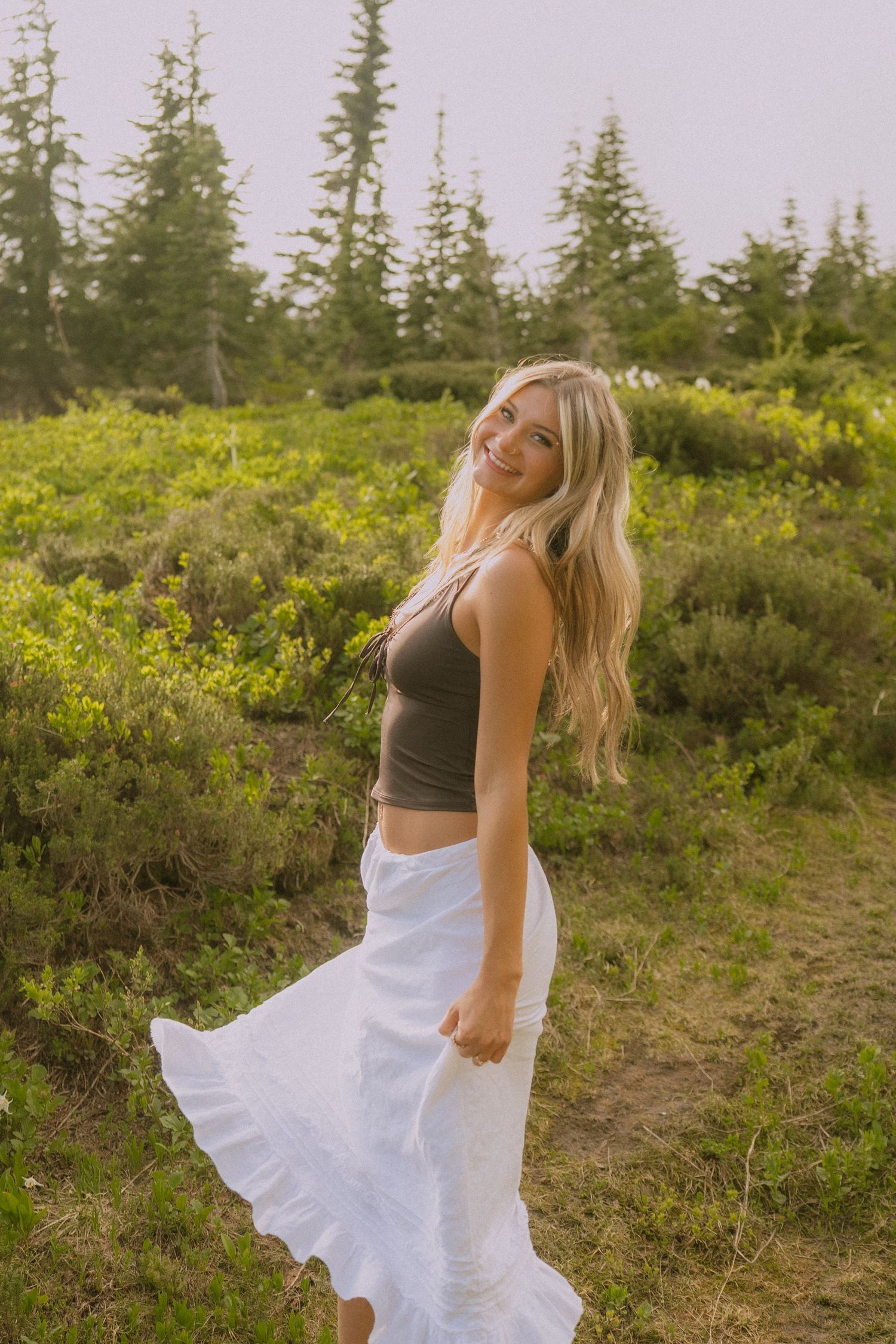 A person in a gray crop top and flowing white skirt twirls in a meadow with evergreen trees and yellow wildflowers.
