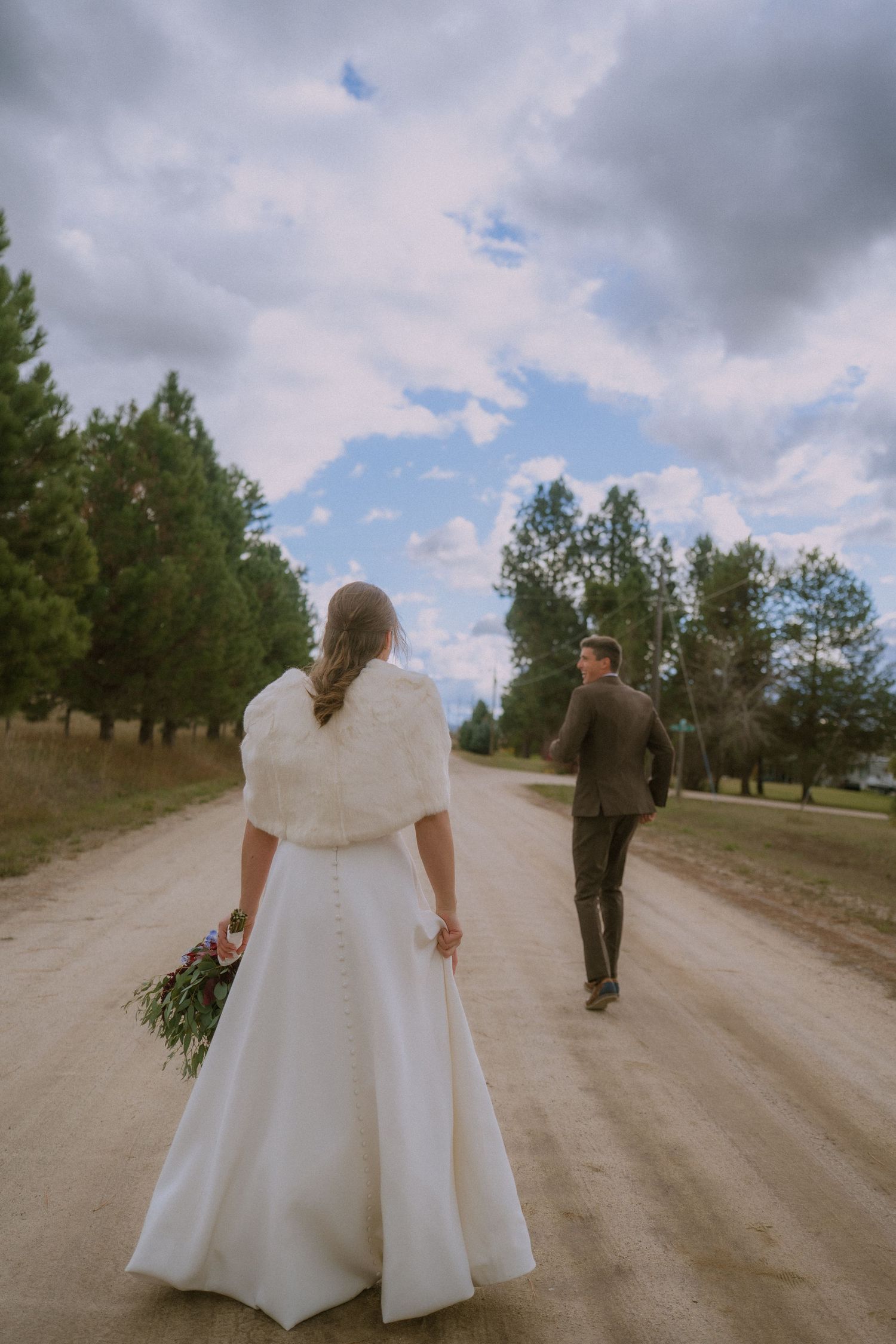 A couple walks down a dirt road together after their wedding ceremony, with the bride wearing a flowing white gown.