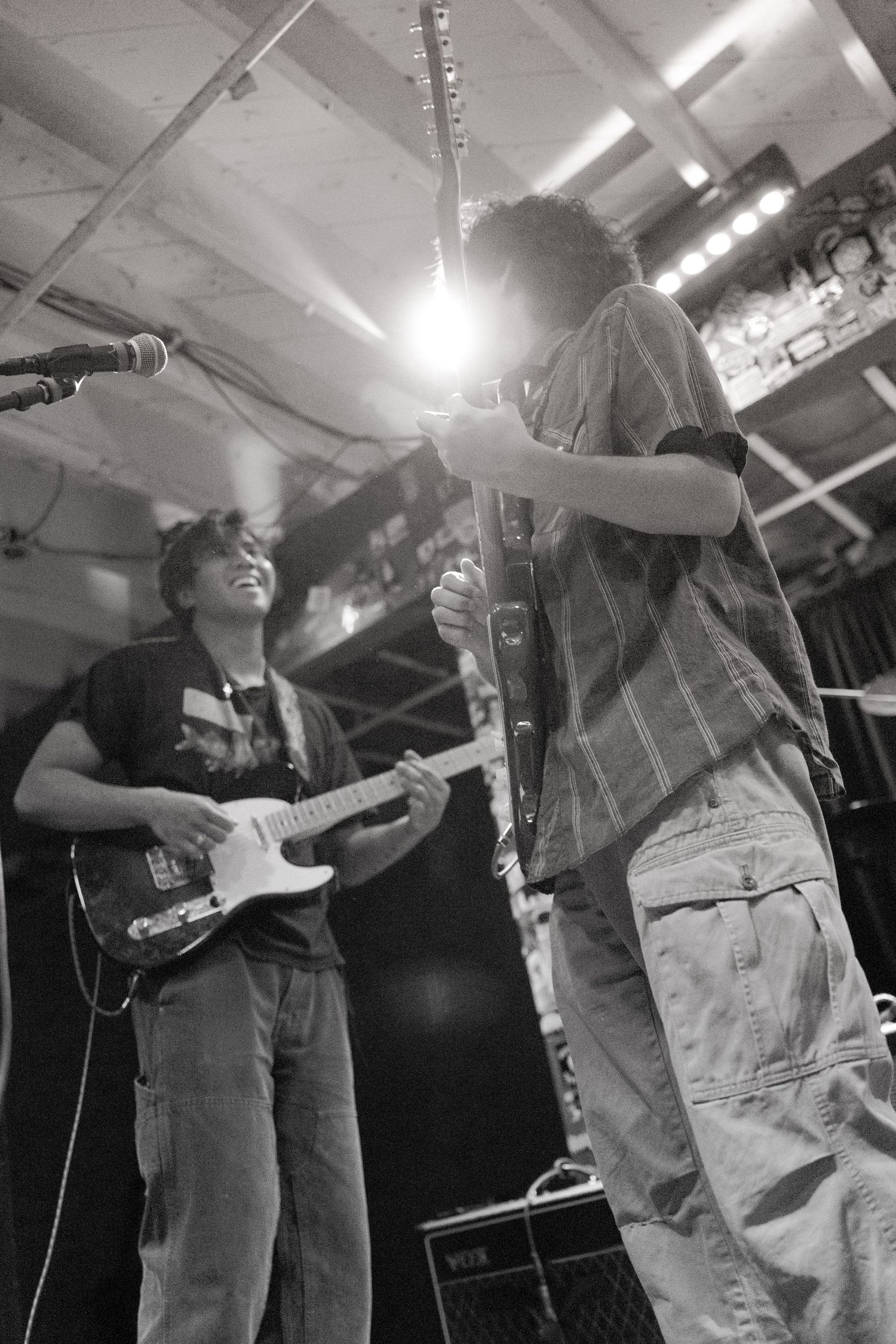 Musicians perform on stage at an intimate venue with electric guitars under bright overhead lighting.
