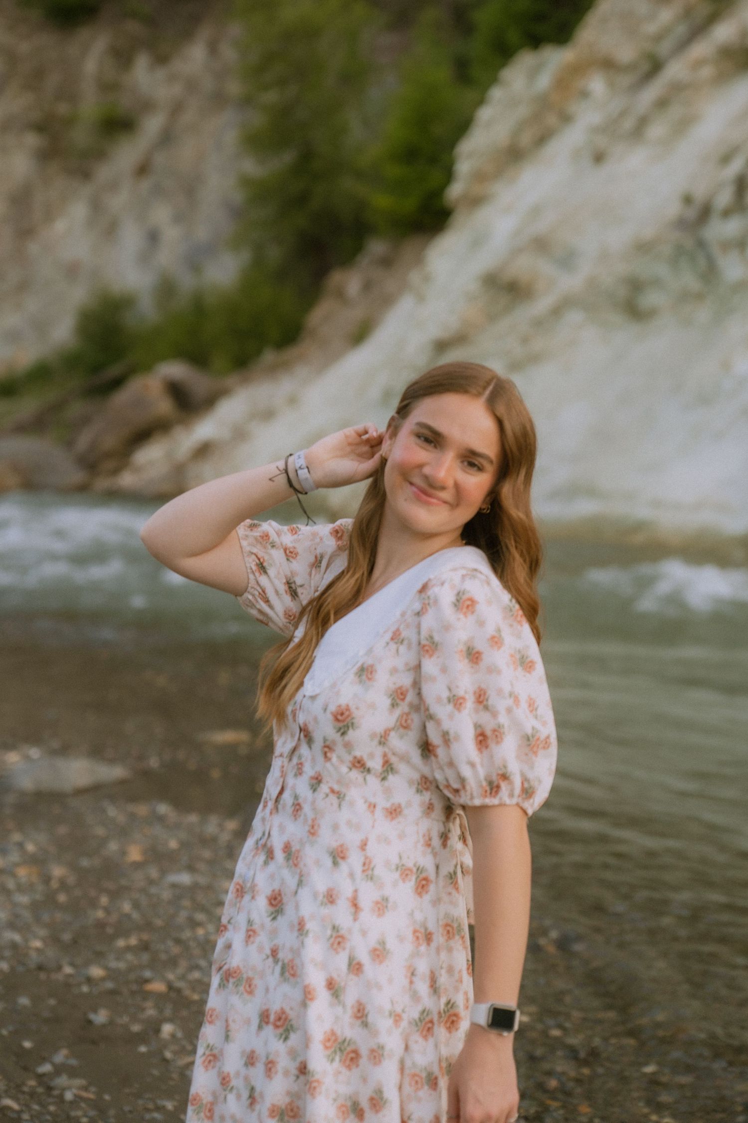 A person in a floral white dress poses near a rocky riverbank with white cliffs in the background.