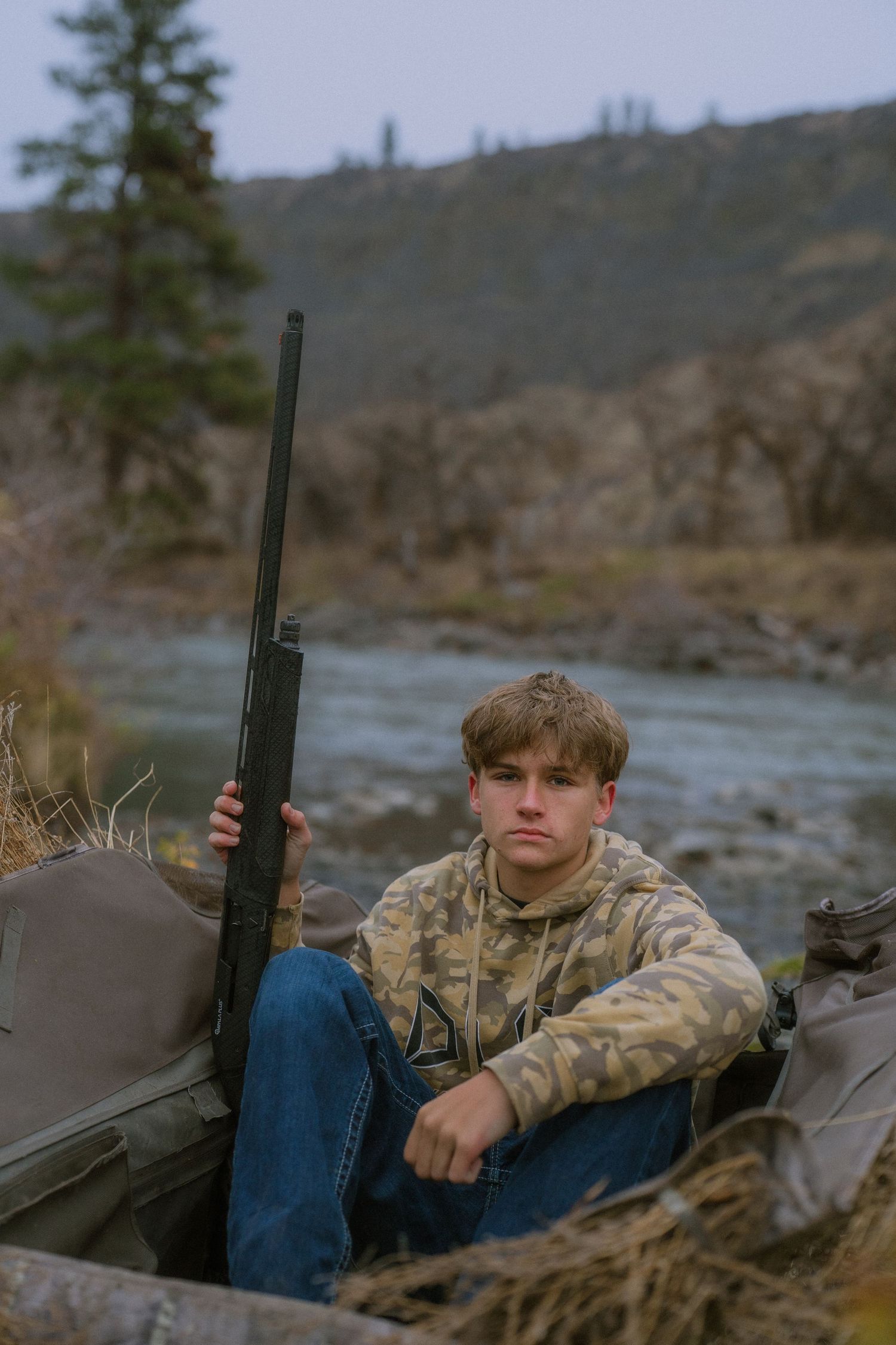 Person in camouflage jacket sits with hunting gear by river in mountainous terrain.