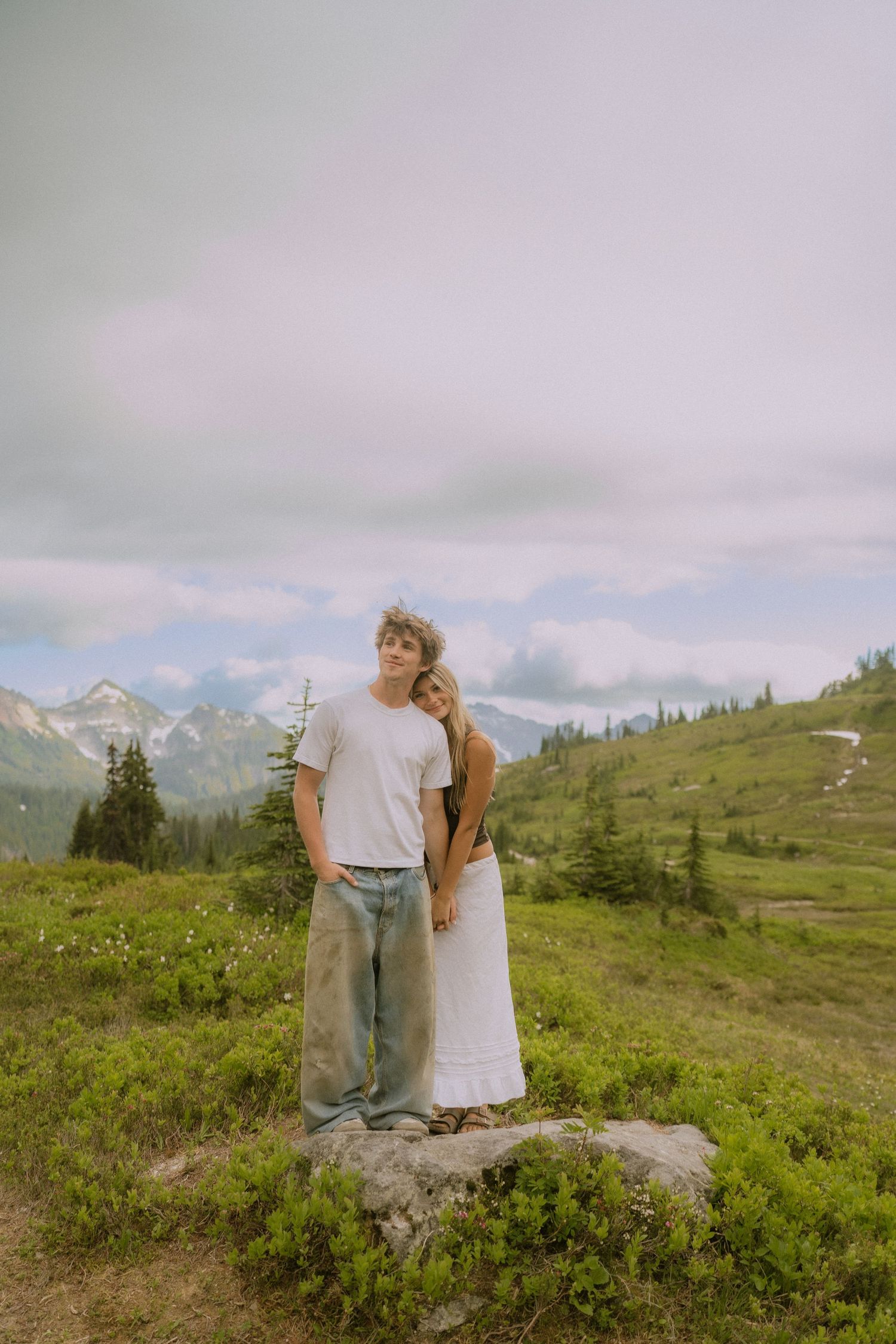 A couple stands together on a rock overlooking a mountainous landscape with wildflowers.