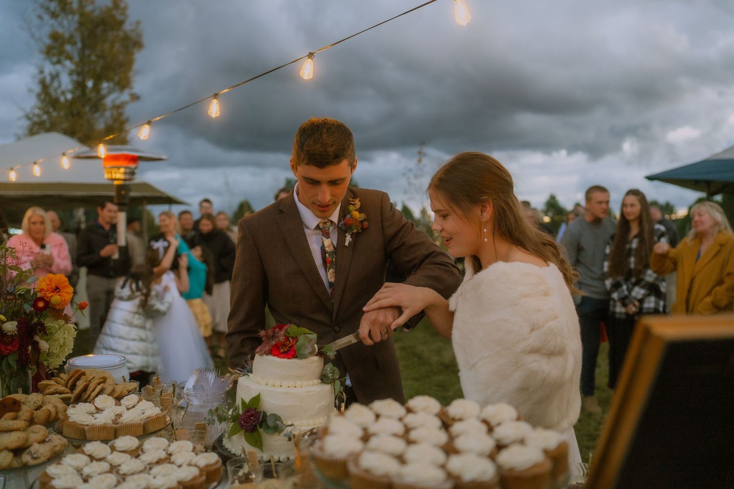 Evening outdoor wedding reception with string lights and dramatic stormy sky overhead.