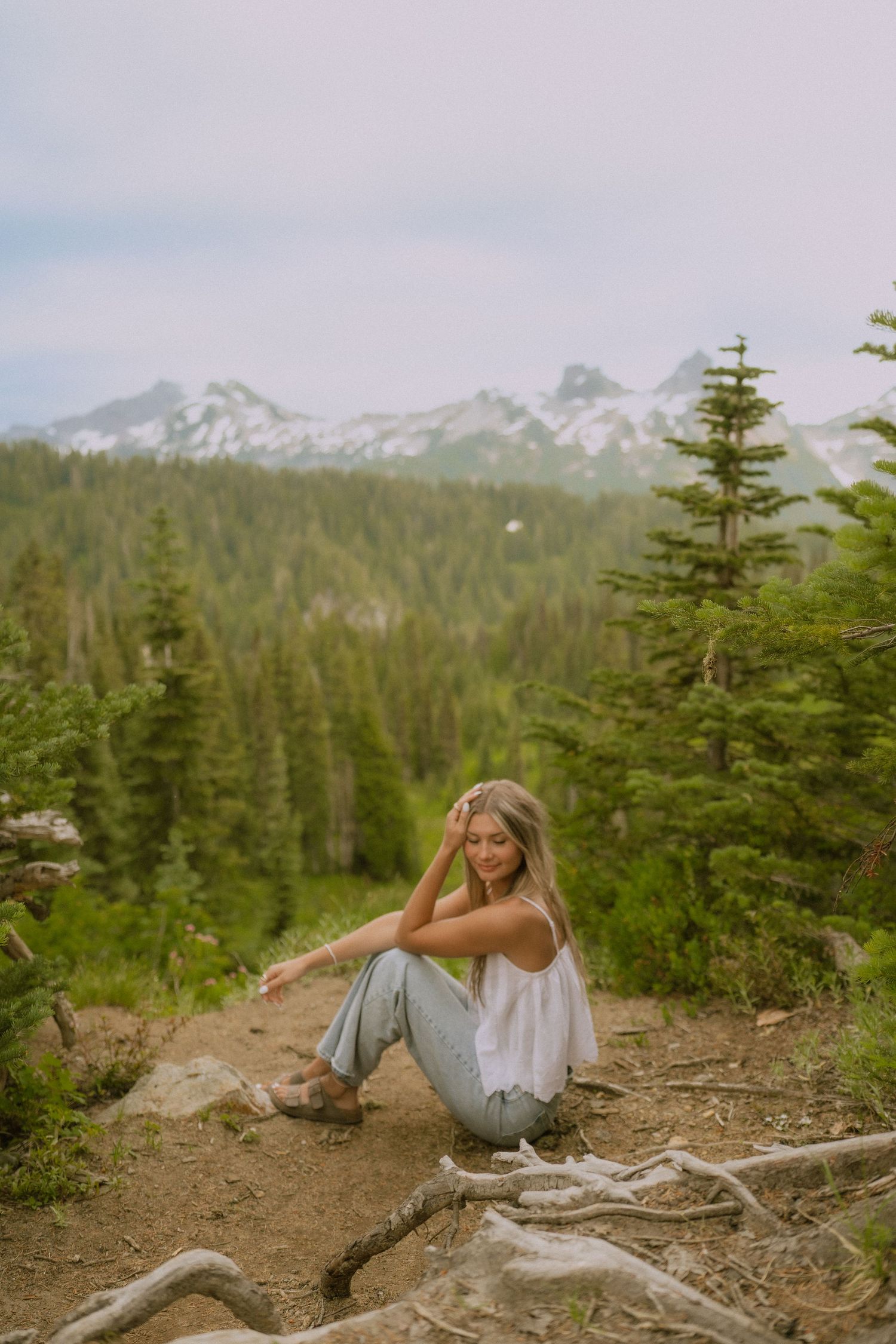 A person in casual attire lounges on a mountain trail surrounded by evergreen trees and snowy peaks in the distance.