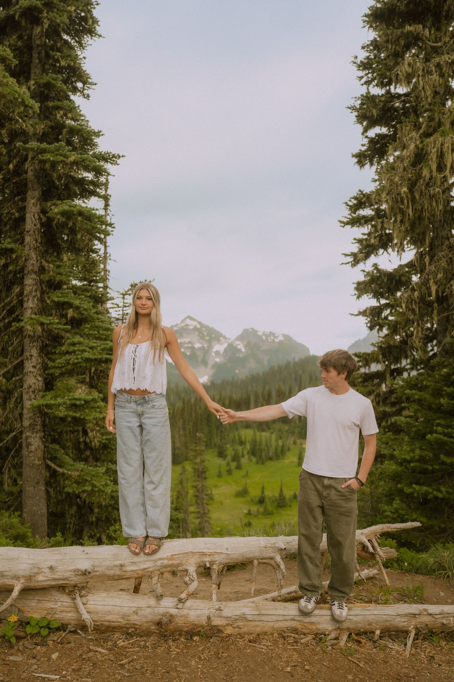 A couple poses playfully on a fallen log with mountain peaks and evergreens in the background.