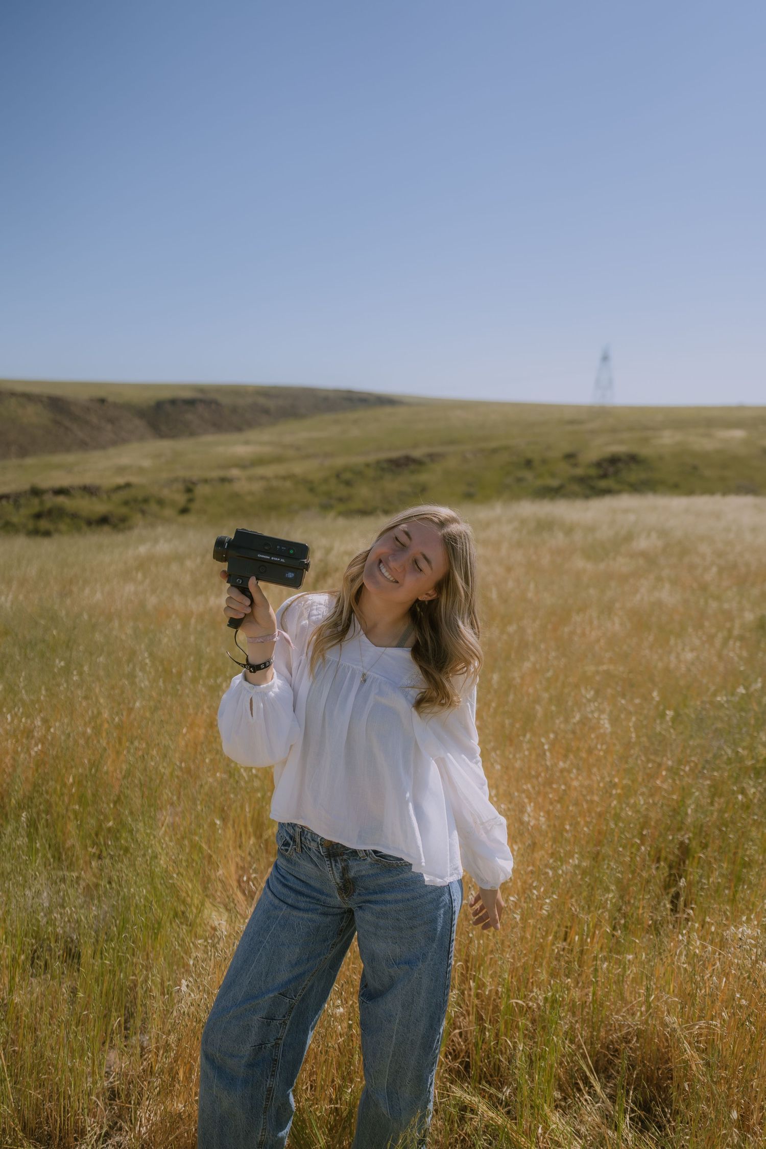 Woman in white blouse and jeans dances freely in golden grassy field under bright summer sky.