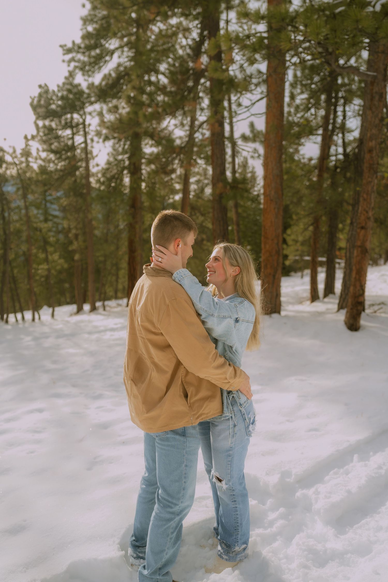 A couple in matching denim and tan jackets playfully embrace in a snowy forest setting during winter.