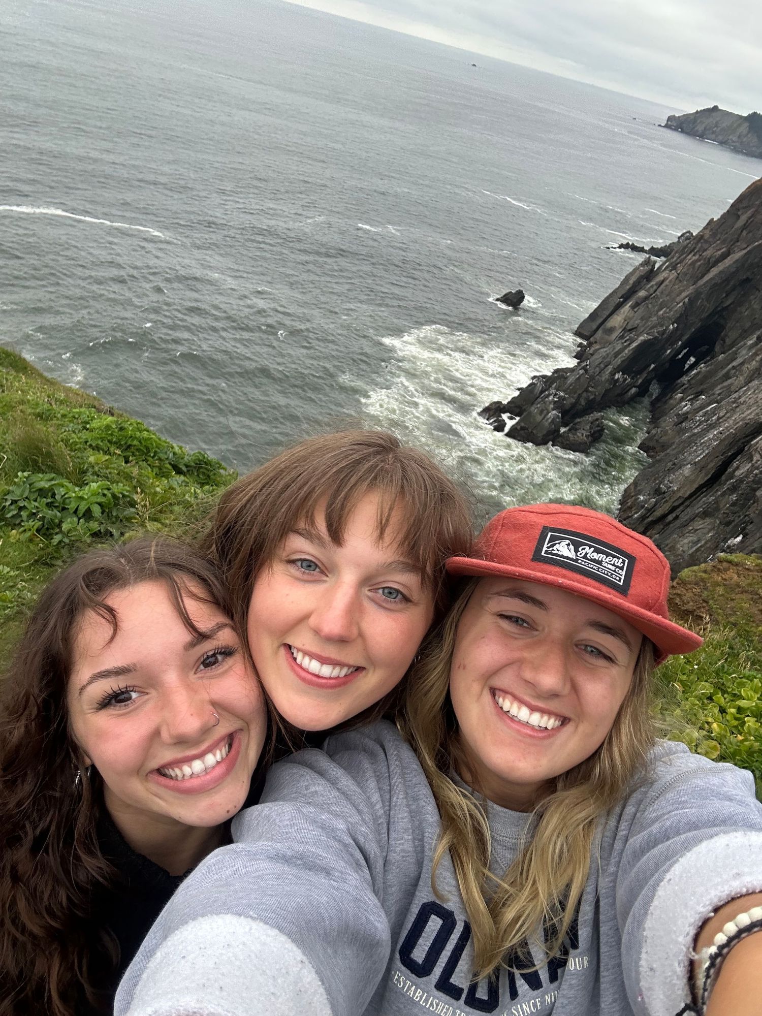 Three friends taking selfies together against a dramatic coastal cliff backdrop overlooking the ocean.