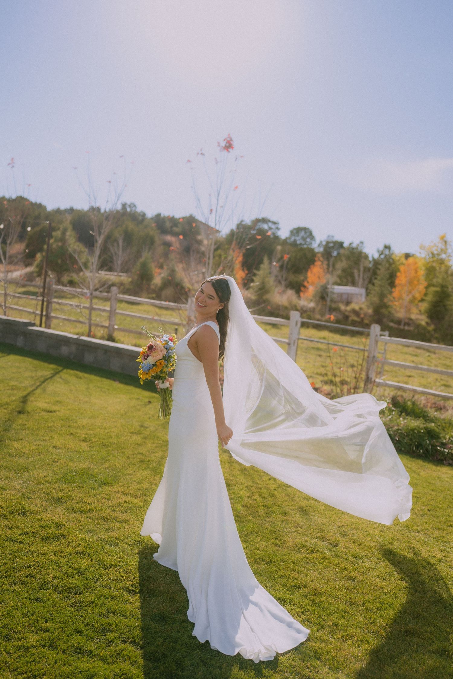 A flowing white wedding dress catches the breeze in a sunny autumn field with fall foliage in the background.