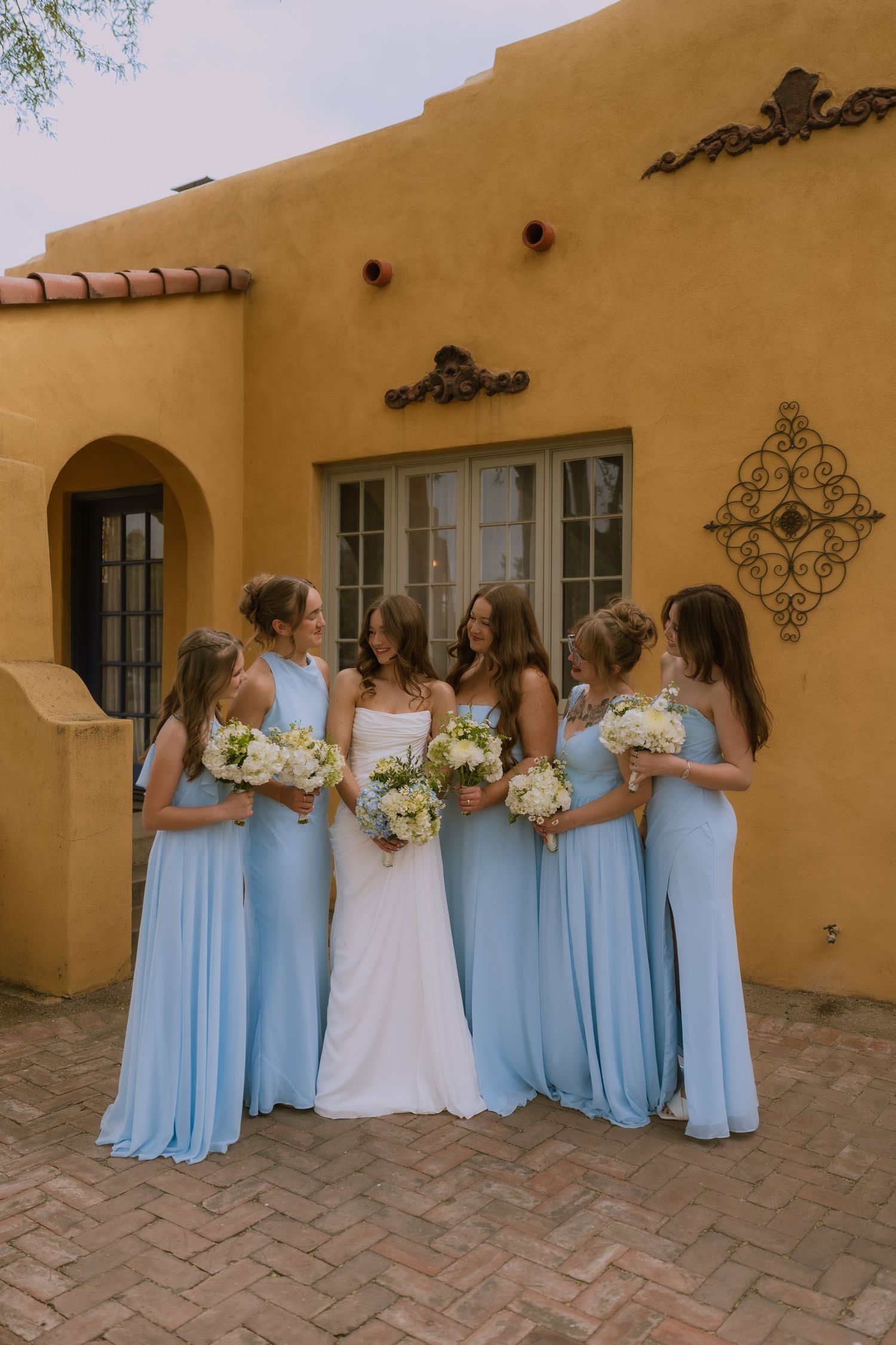 Bridesmaids in light blue dresses stand with bride against adobe wall at outdoor wedding venue.