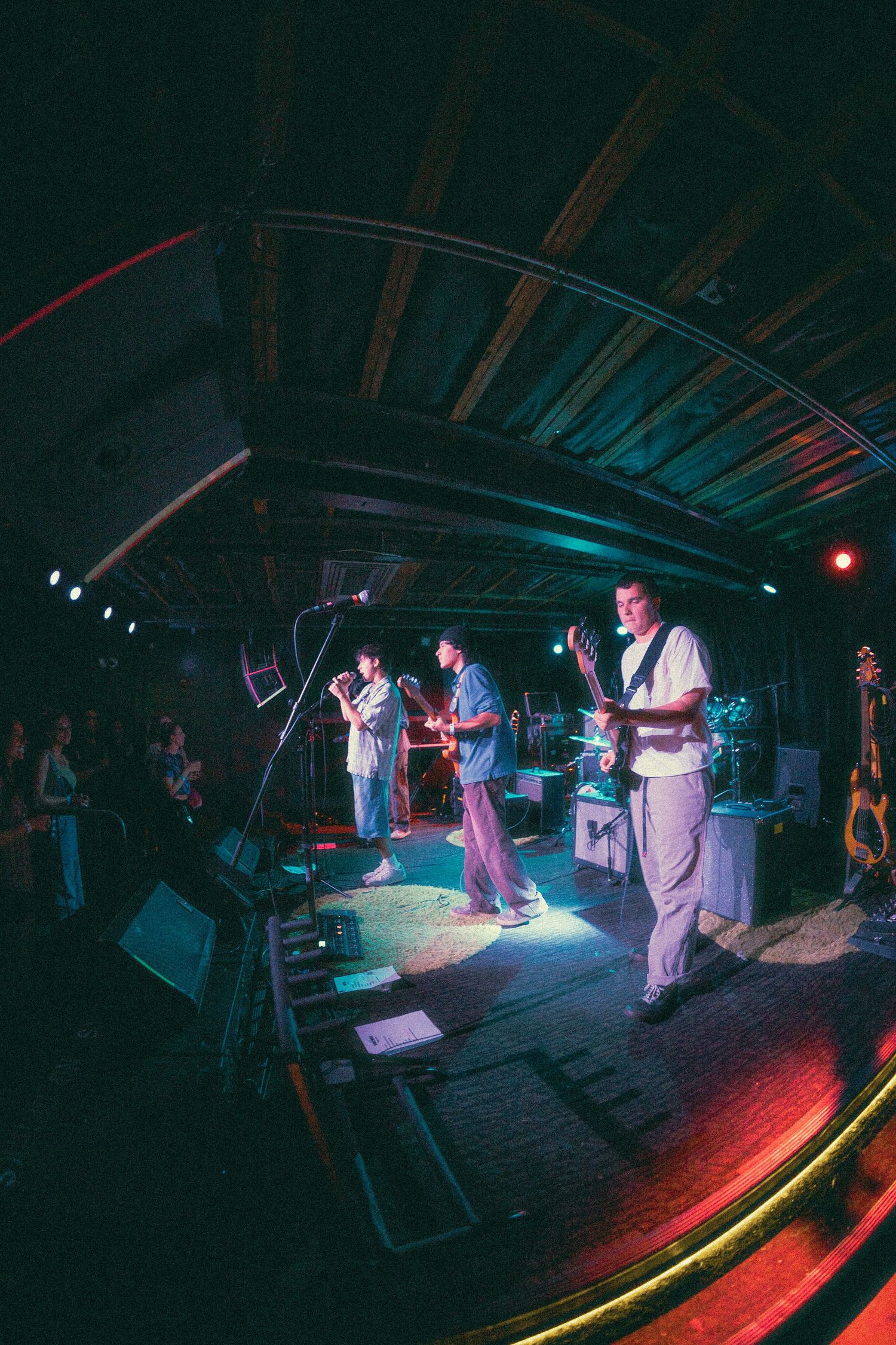 Wide-angle shot of a band playing in a dark venue with colorful stage lighting and equipment setup.