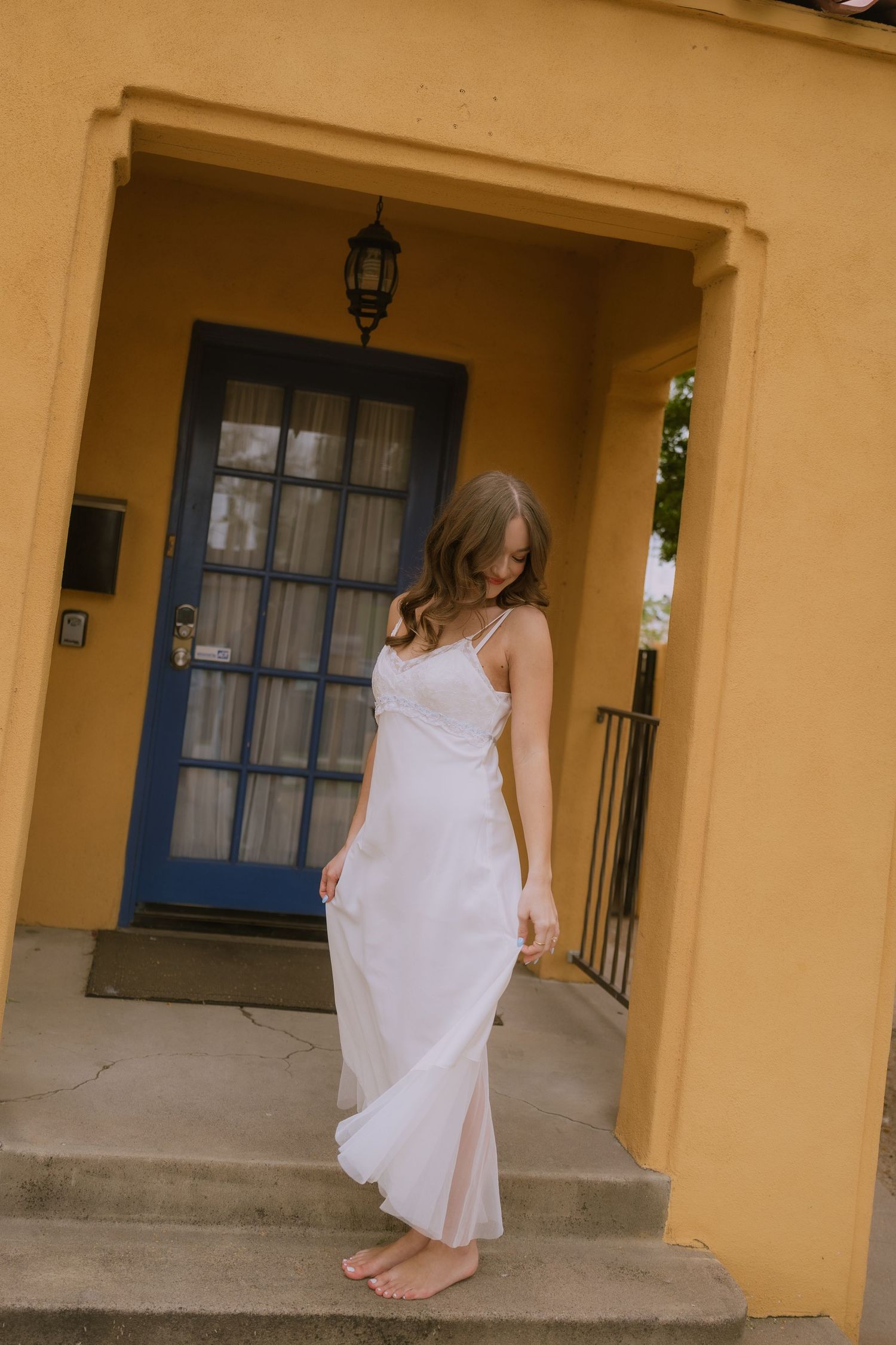 A person in a flowing white dress walks along a covered porch with yellow walls and blue doors.