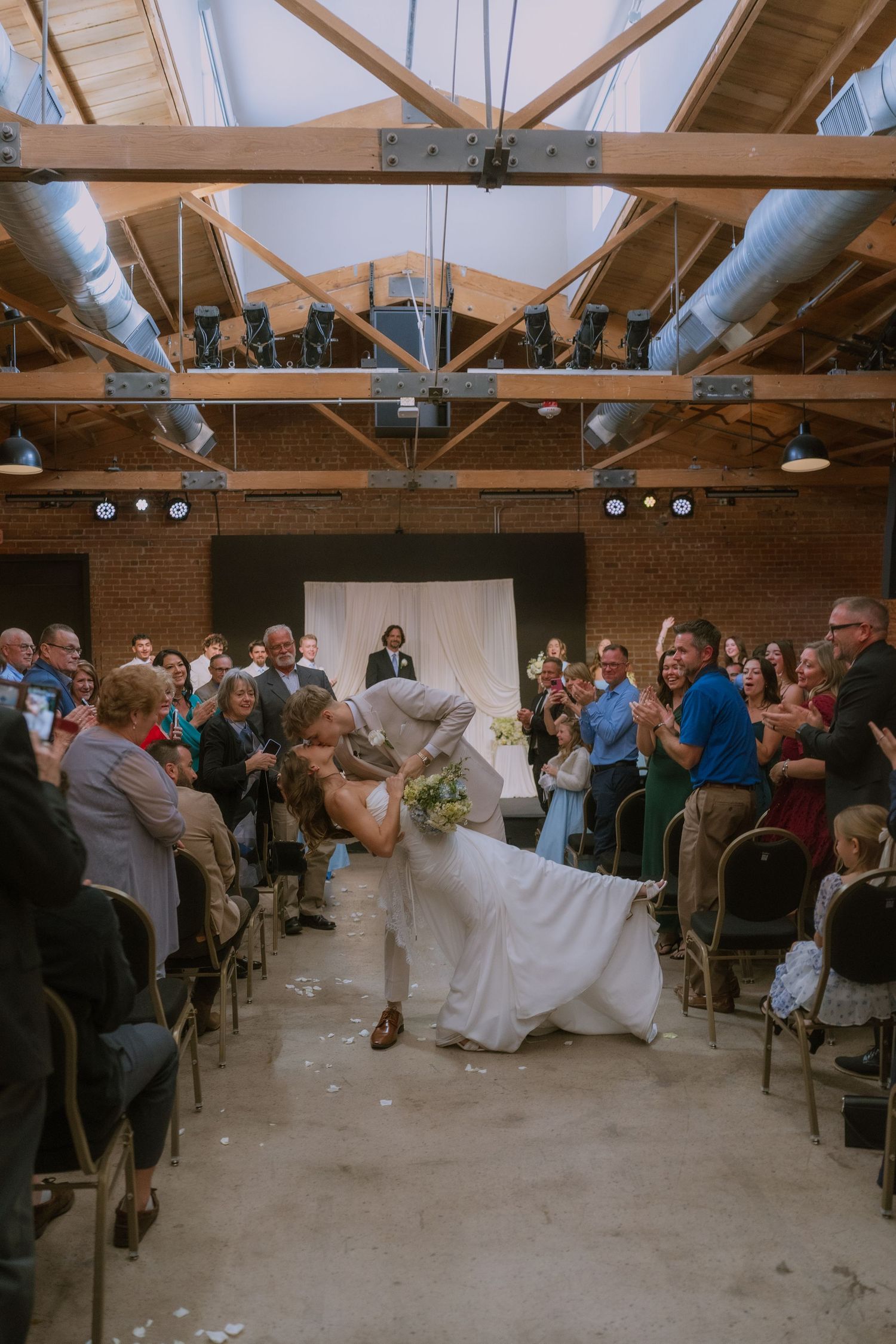 Wedding guests gather in a rustic venue with exposed wooden beams and industrial lighting during a reception celebration.