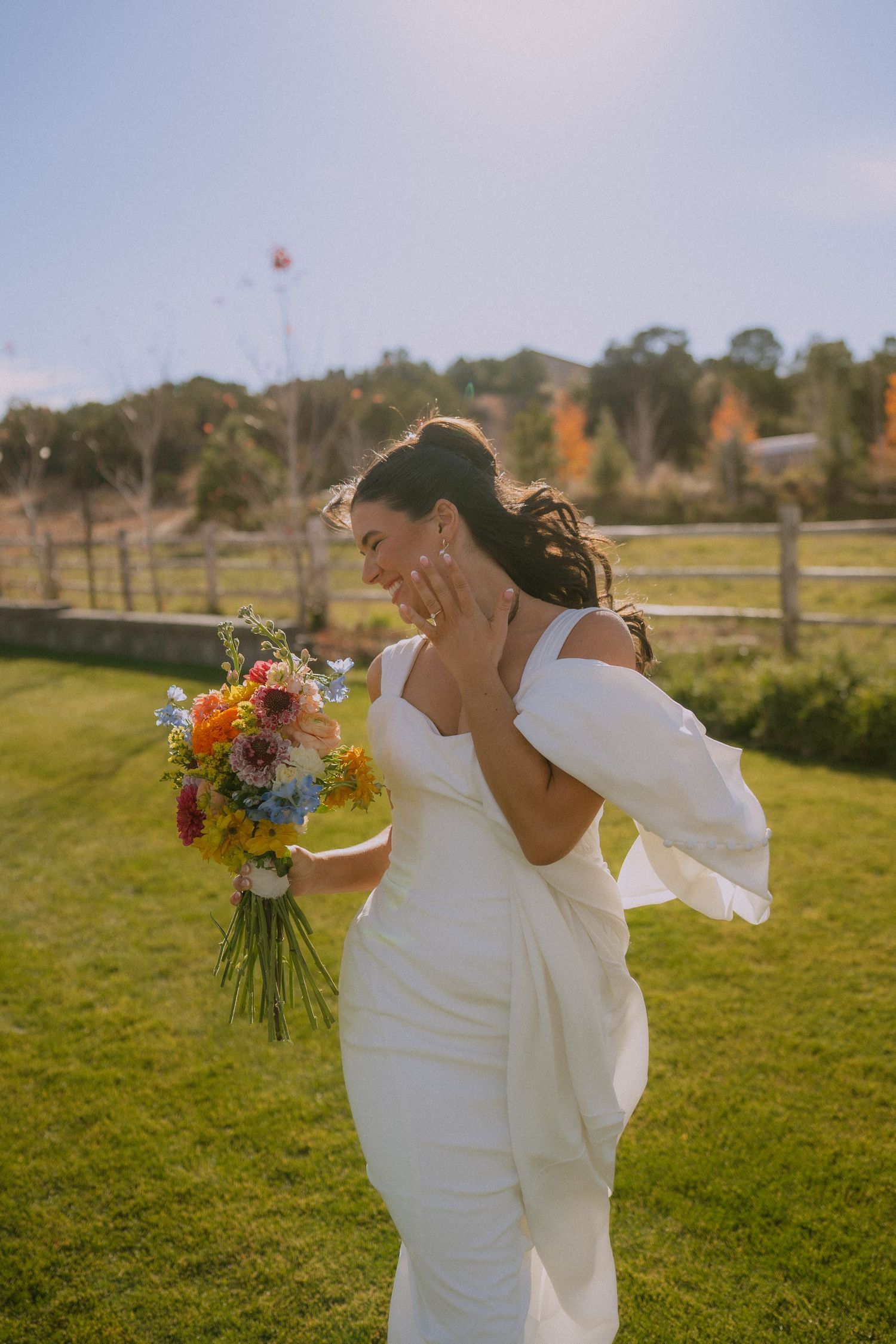 Two brides share a joyful moment in a sunny outdoor field with colorful bouquets.