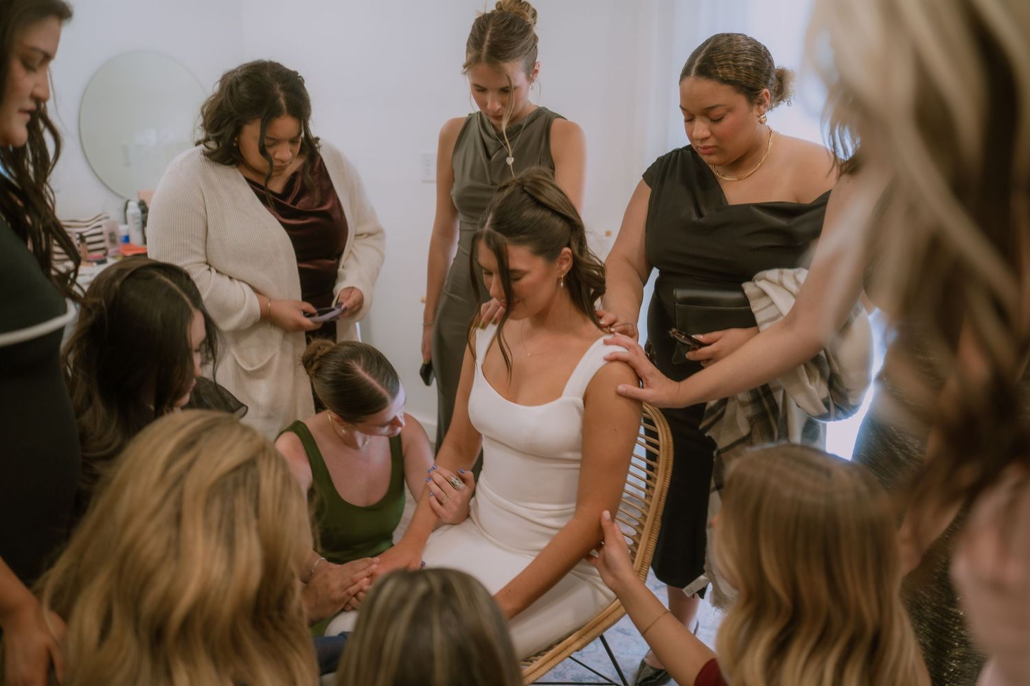 A bride in a white dress sits surrounded by her bridal party during wedding preparations in warm lighting.