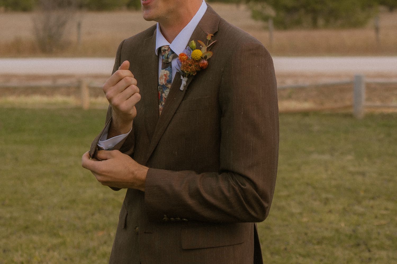A person in a brown suit with a boutonniere stands outdoors in a grassy field during golden hour.