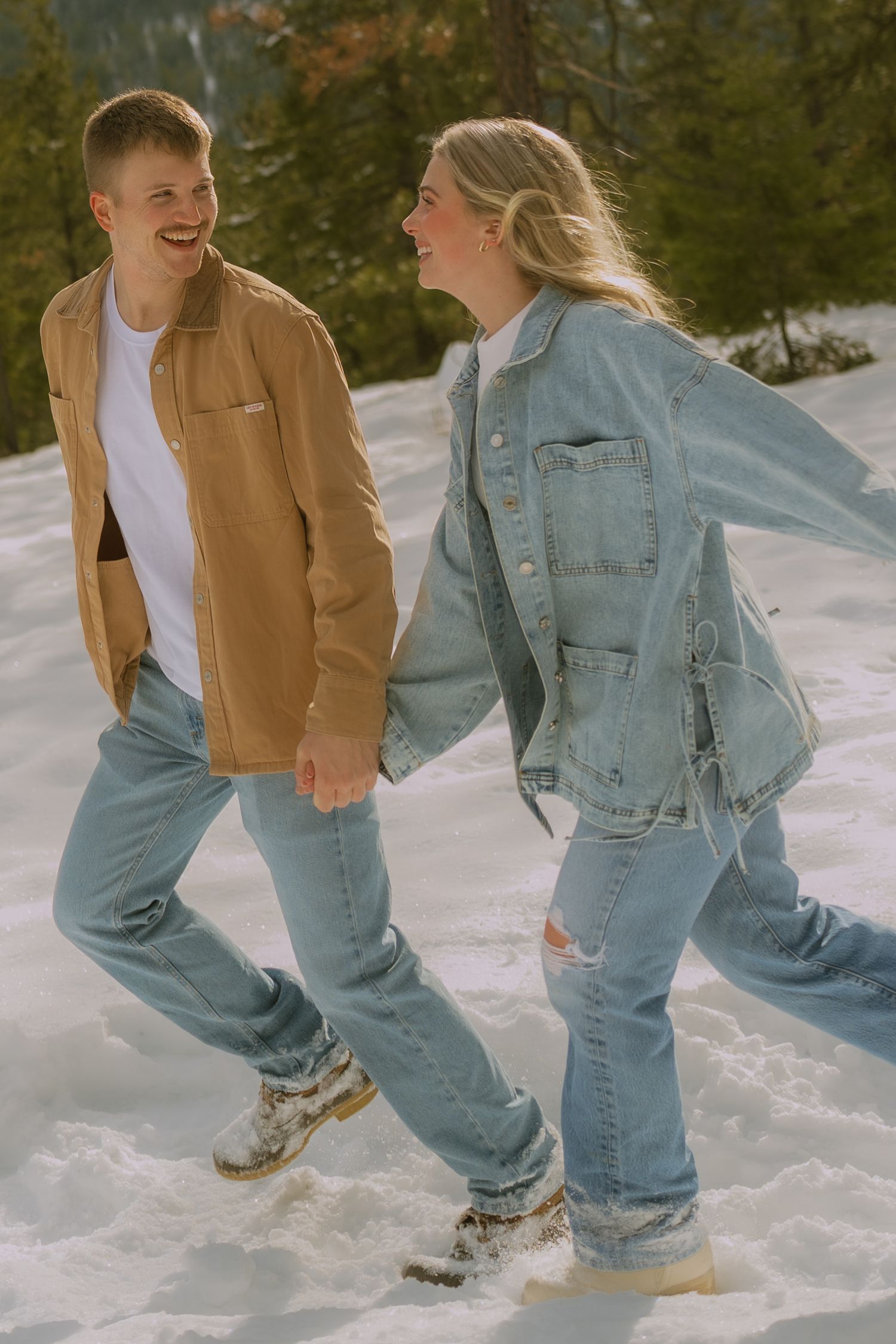 A couple walking through snowy terrain wearing matching denim and jackets while sharing a laugh in winter sunlight.