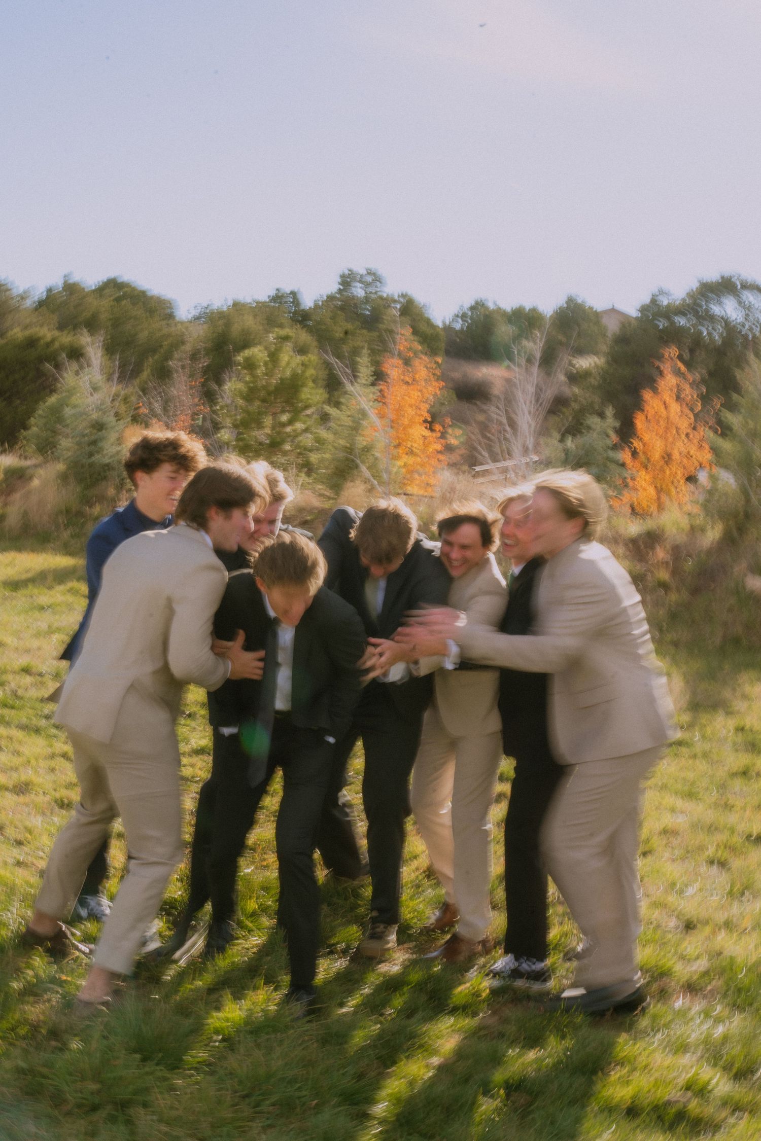 A group of people huddled together laughing in a sunny field with autumn trees in the background.