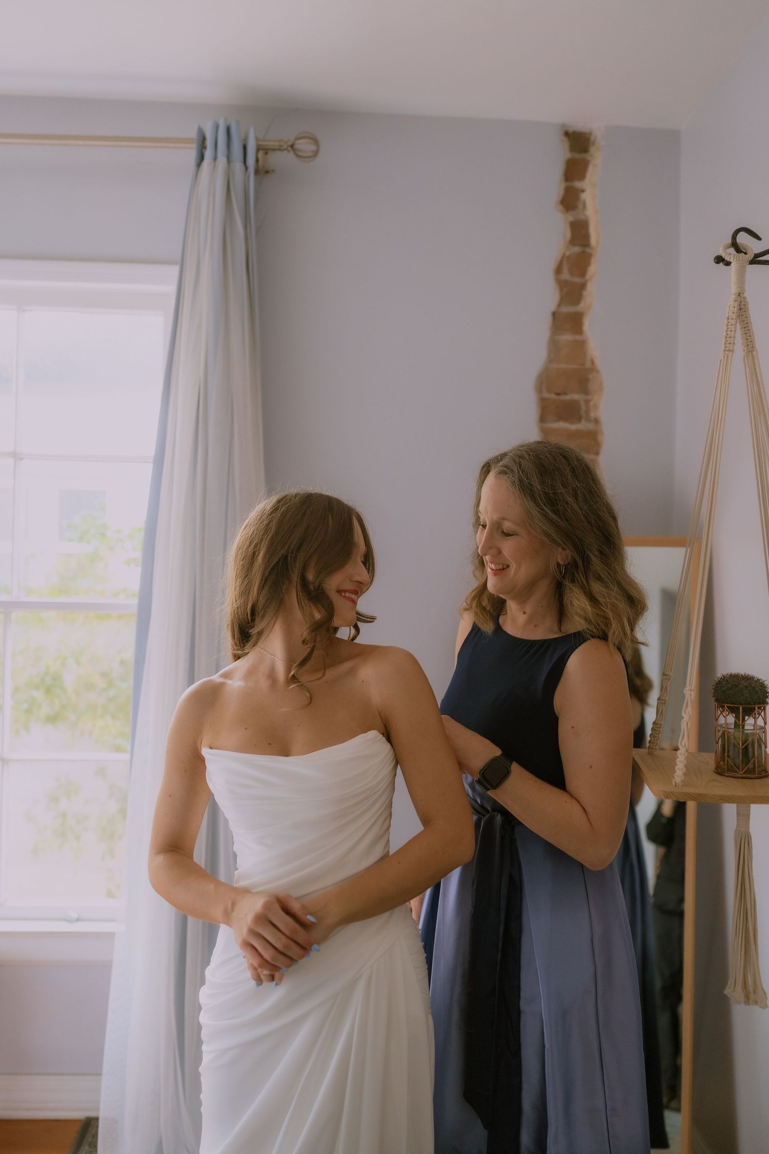 Two women share an intimate moment while getting ready in a sunlit room with white curtains and a wedding dress.