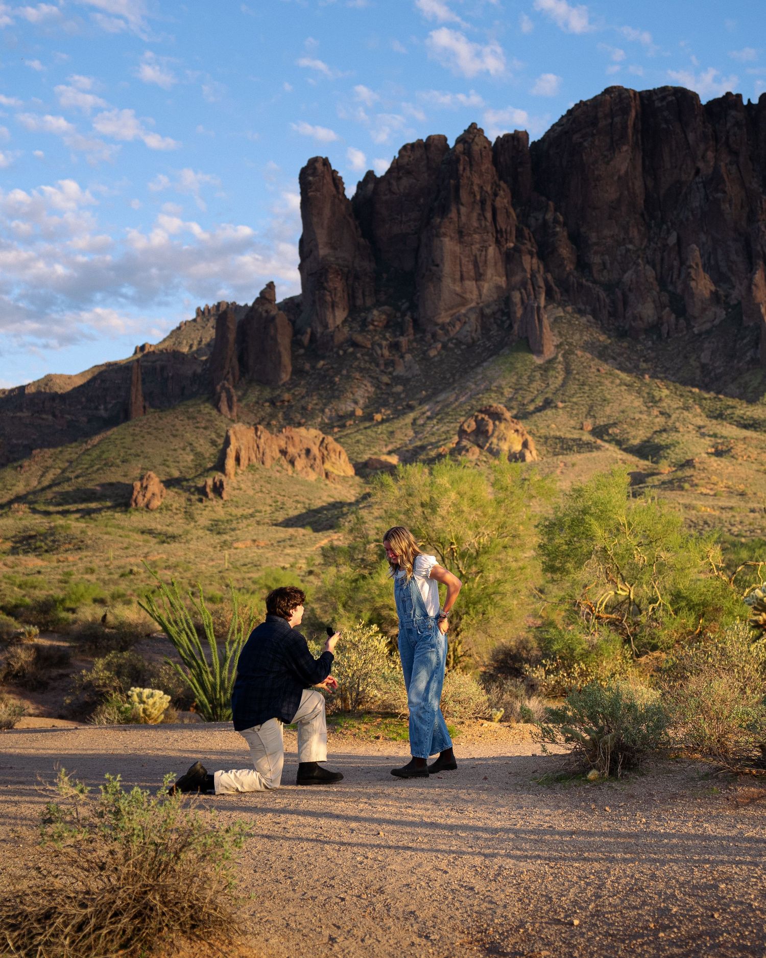 Two people share a special moment on a desert trail with dramatic Superstition Mountains rising in the background at sunset.