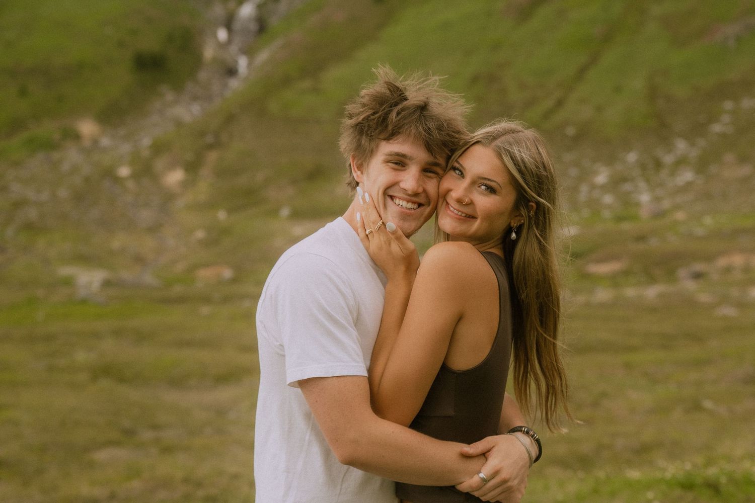 A romantic couple embraces and laughs together on a grassy hillside during a sunny afternoon.
