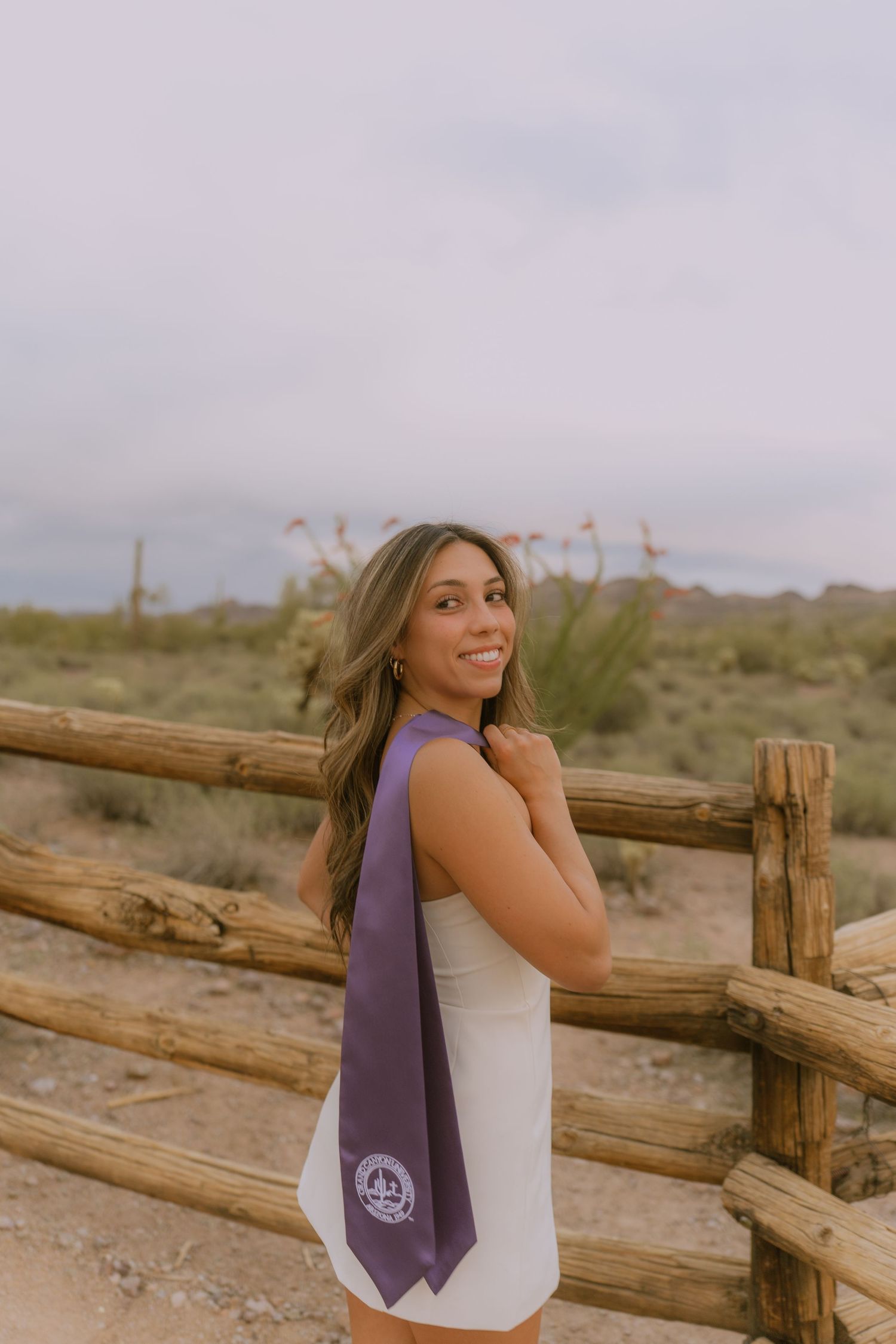 A person wearing a white dress and purple sash stands by a wooden fence in a desert landscape at sunset.