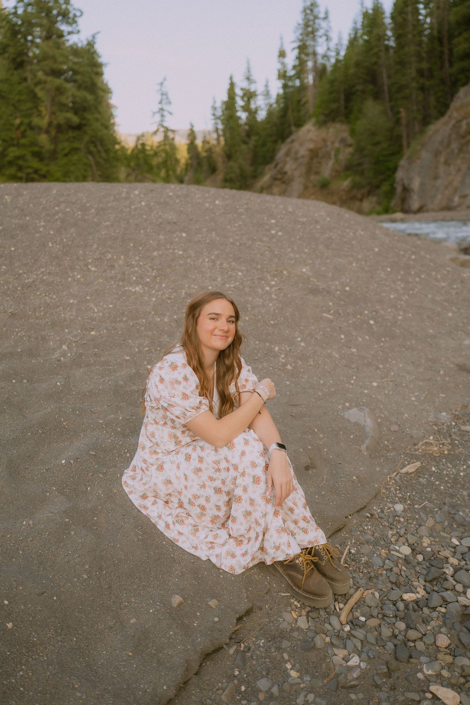 A floral dress contrasts against rocky terrain near a river with pine trees in the background.