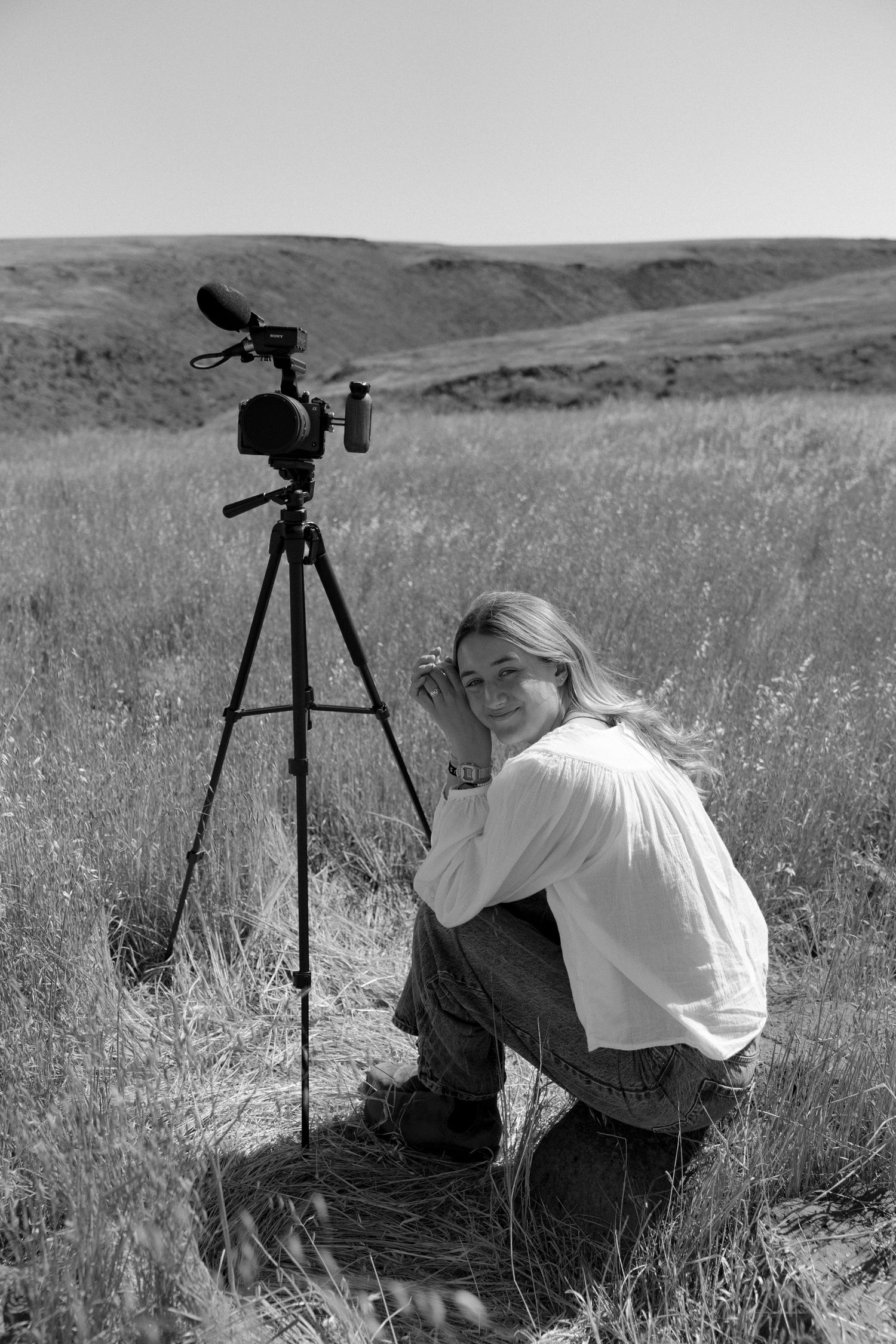 Two photographers with tripod-mounted cameras work in a grassy meadow against rolling hills in black and white.