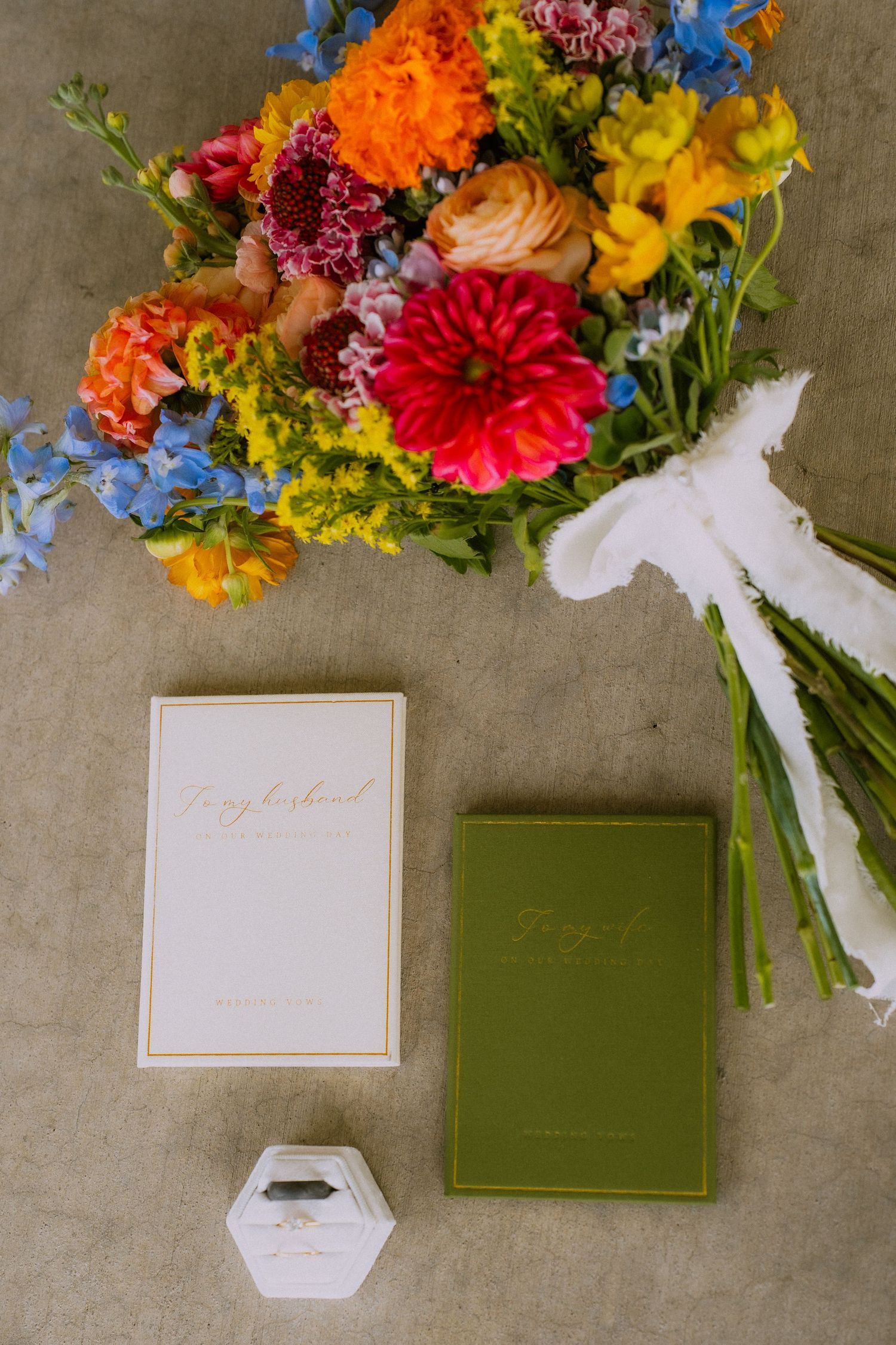 Colorful wedding bouquet with dahlias and wildflowers next to white and green notebooks and a small ring box.