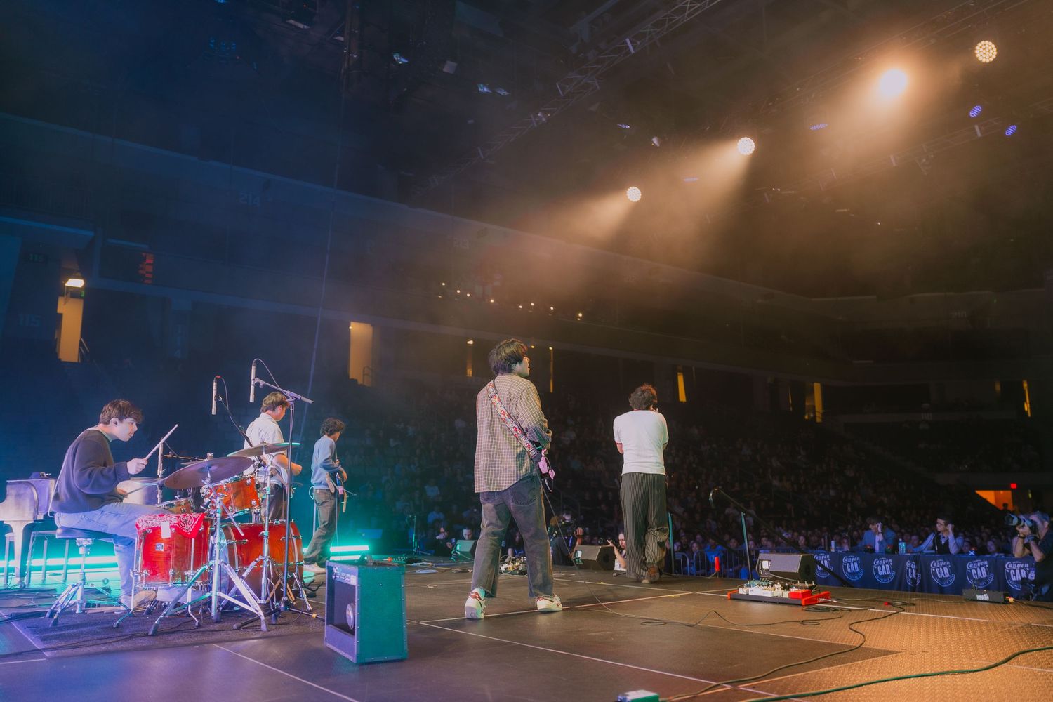 Musicians perform on stage during a concert with dramatic lighting and a crowd visible in the background.